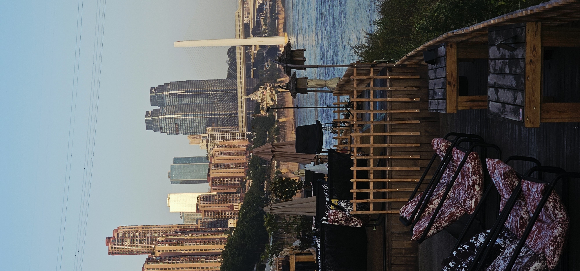 The image depicts an outdoor seating area overlooking a river and a city skyline. The foreground shows a wooden deck with several lounge chairs, some with red and white patterned cushions. Two wooden tables with a weathered finish are also visible. Beyond the deck, a low wooden fence separates the seating area from the riverbank. In the middle ground, there are several outdoor umbrellas and chairs, suggesting a restaurant or cafe. The background features a wide river with a prominent cable-stayed bridge in the distance. The cityscape includes numerous tall buildings, with a cluster of modern, glass-fronted skyscrapers dominating the view. The sky is clear and pale blue, indicating daytime. The scene is located in the Nan'an District of China, with the architecture and urban layout characteristic of a riverside city in the region. There are no people visible in the image.