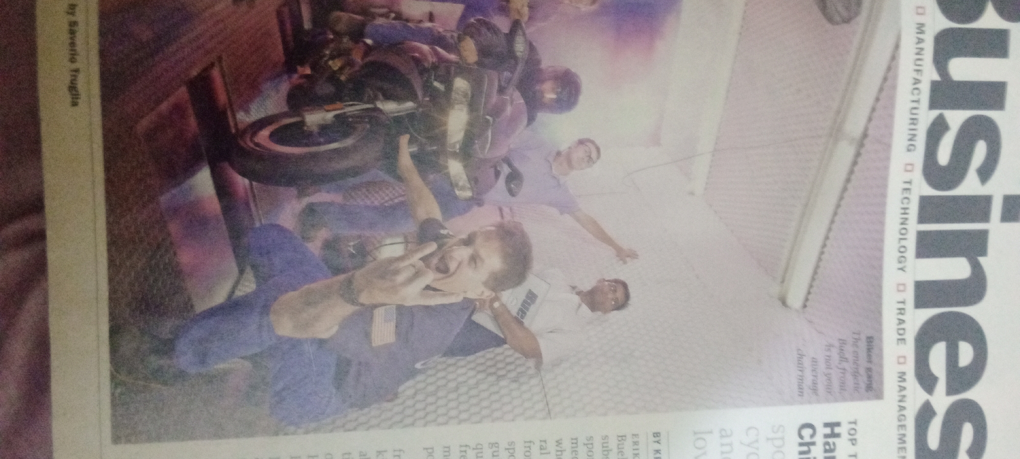The image shows a newspaper spread with a motorcycle and several individuals. A person in the foreground is wearing a blue shirt with an American flag patch and making a "rock on" hand gesture. Behind them, a motorcycle is partially visible. Several other individuals are in the background, some appearing to be looking at the motorcycle or the camera. The text on the right side of the image includes the word "Business" in large font and other smaller text that appears to be article headings and bylines. The image also includes text on the left side with a byline and what appears to be an article about motorcycles. The environment appears to be indoors, possibly a studio or a staged setting, given the lighting and the background. The image does not contain any information that can identify its location in the city of Jalingo, Nigeria.