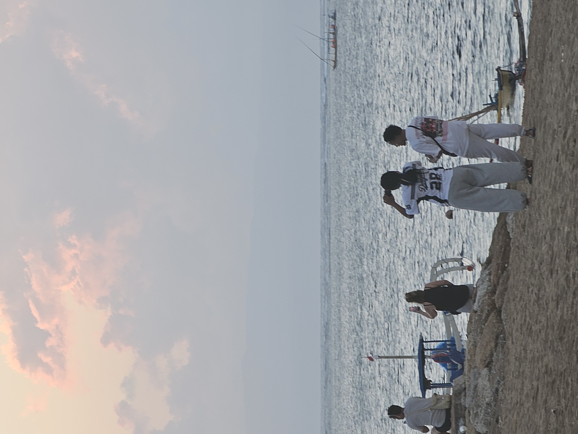 At dusk on a beach in Denpasar, Indonesia, four individuals are positioned near the water's edge. The sky is filled with soft, pink-tinged clouds. The sea is calm, with gentle waves lapping the shore. In the distance, a boat with two masts sails across the horizon. A smaller boat, possibly a fishing boat, is moored closer to shore, near a blue canopy structure.
In the foreground, a person wearing a white t-shirt and light-colored pants sits facing away from the camera, a bag slung over their shoulder. To their right, another person, dressed in a black tank top and light-colored shorts, stands and holds up a mobile phone, appearing to take a photograph. Further to the right, two individuals stand together, facing the water. One, wearing a white t-shirt with graphics and light-colored pants, holds a phone up as if taking a picture. The other, in a white t-shirt and gray pants, is looking away from the camera. The ground is a mix of sand and rocks.