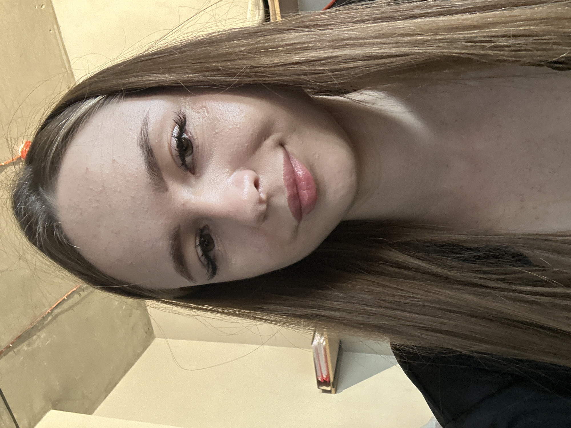 A young woman with long, straight brown hair is shown in a close-up, waist-up portrait. She is looking down and slightly to the left, with a subtle smile on her lips. Her eyes are brown, and she is wearing false eyelashes. The background is blurred, but appears to be an indoor setting with a light-colored wall and a concrete ceiling with visible pipes. There is a shelf with some books or papers on it to the left of the woman. She is wearing a black garment.