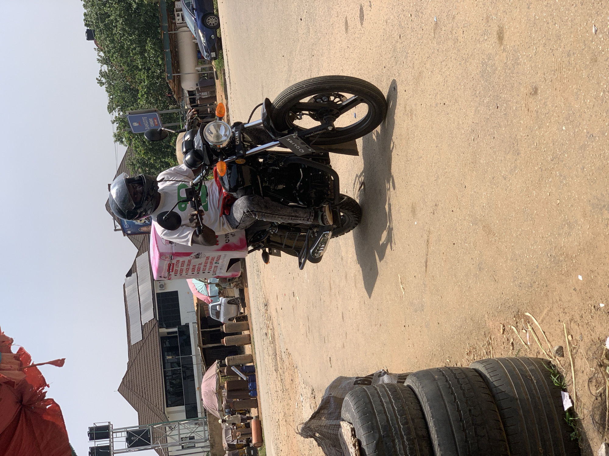 The image depicts a street scene in Abuja, Nigeria, likely in a commercial or mixed-use area. A person wearing a black helmet and a white t-shirt with green lettering is riding a black motorcycle. The motorcycle has a pink delivery box mounted on the back, with visible text that includes "BODY LOTION," "BODY SCRUB," "FACE CREAMS," "SERUM," and "BODY OIL." The rider is wearing gray denim jeans and appears to be in motion.
In the background, there are buildings with tiled roofs, some featuring solar panels. A white SUV is parked near a structure with a red umbrella. Several tall, cylindrical objects, possibly gas cylinders, are arranged along the roadside, with a few smaller blue containers nearby. A sign that reads "DANGOTE CEMENT" is visible on a blue post. To the left of the motorcycle, a stack of used tires is piled on the ground, partially covered by a dark net bag. The road surface is unpaved and dusty, indicative of a typical Nigerian urban environment. The sky is clear and bright, suggesting a sunny day.