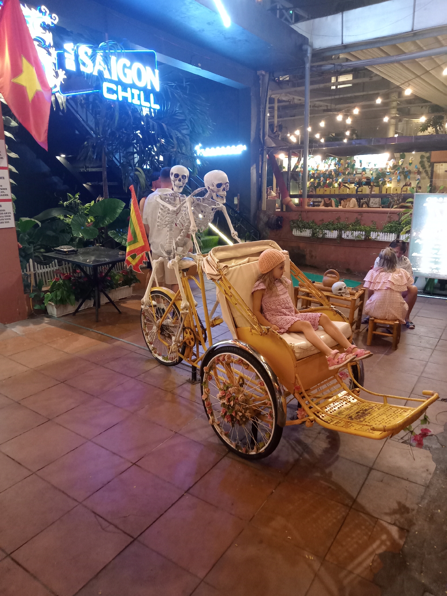This image captures a lively night scene at an outdoor or semi-outdoor establishment, likely a cafe or restaurant, in Nha Trang, Vietnam. The focal point is a young child, possibly a girl, with blonde hair and a peach-colored sun hat, sitting in the passenger seat of a decorative, bright yellow cycloshow. She is wearing a patterned pink dress and pink shoes, and appears calm.
Behind the cycloshow, two life-sized skeleton figures are positioned as if riding the back of the cycloshow, adding a quirky or Halloween-themed touch to the decor. A Vietnamese flag, with its red field and yellow star, hangs prominently on the left. In the background, other patrons are visible, with two individuals seated at a low table to the right of the cycloshow, and more people seen further back in a brightly lit area with string lights. The setting features lush green plants and small potted flowers, indicating an inviting ambiance.
The time of day is clearly night, as evidenced by the illuminated neon sign reading "SAIGON CHILL" in bright blue letters, and various overhead lights that cast a warm glow on the scene. The ground is paved with brown square tiles.
Visible text includes the prominent "SAIGON CHILL" sign. Partially visible text on a white sign to the left includes "8%", "MARKE", "2026", "DD AN 8%", "LIL UPON", "ATE YOUR", and "NK YOU", possibly from a menu or promotional material. The overall impression is one of a vibrant, decorated tourist spot or local eatery.