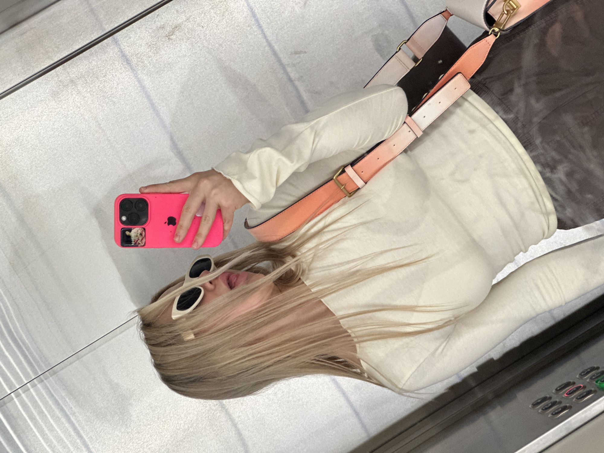A woman with long, blonde hair is taking a selfie in an elevator. She is wearing a light-colored, long-sleeved shirt and white sunglasses. She is holding a bright pink iPhone with multiple lenses in her right hand. A handbag with a light pink and peach strap is slung over her shoulder. The elevator interior appears to be made of brushed metal with visible buttons. The image appears to be taken in Budva, Montenegro.