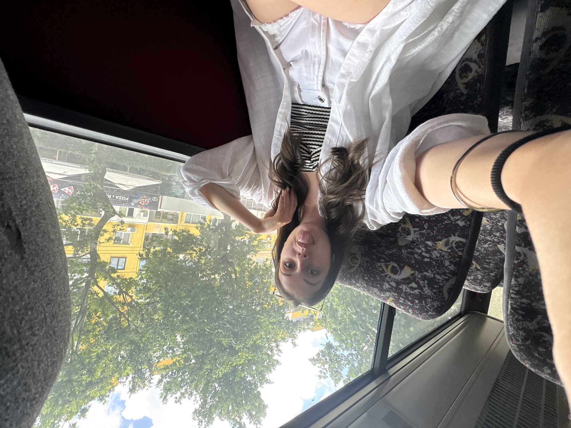 A young woman is taking a selfie while sitting on a bus in Burgas, Bulgaria. The image is taken from a slightly high angle, looking down at the woman. She is wearing a white open shirt over a black and white striped top. Her hair is long and brown, and she is sticking her tongue out playfully at the camera. She is also wearing glasses and a thin silver bracelet on her left wrist. The interior of the bus is visible, with patterned seats and a metal railing. Outside the window, trees and a yellow building can be seen, suggesting it is daytime and the weather is fair. The bus appears to be moving through an urban or suburban area. There is visible text on a sign in the distance, which appears to be advertising "PIZZA & ANGELO'S."