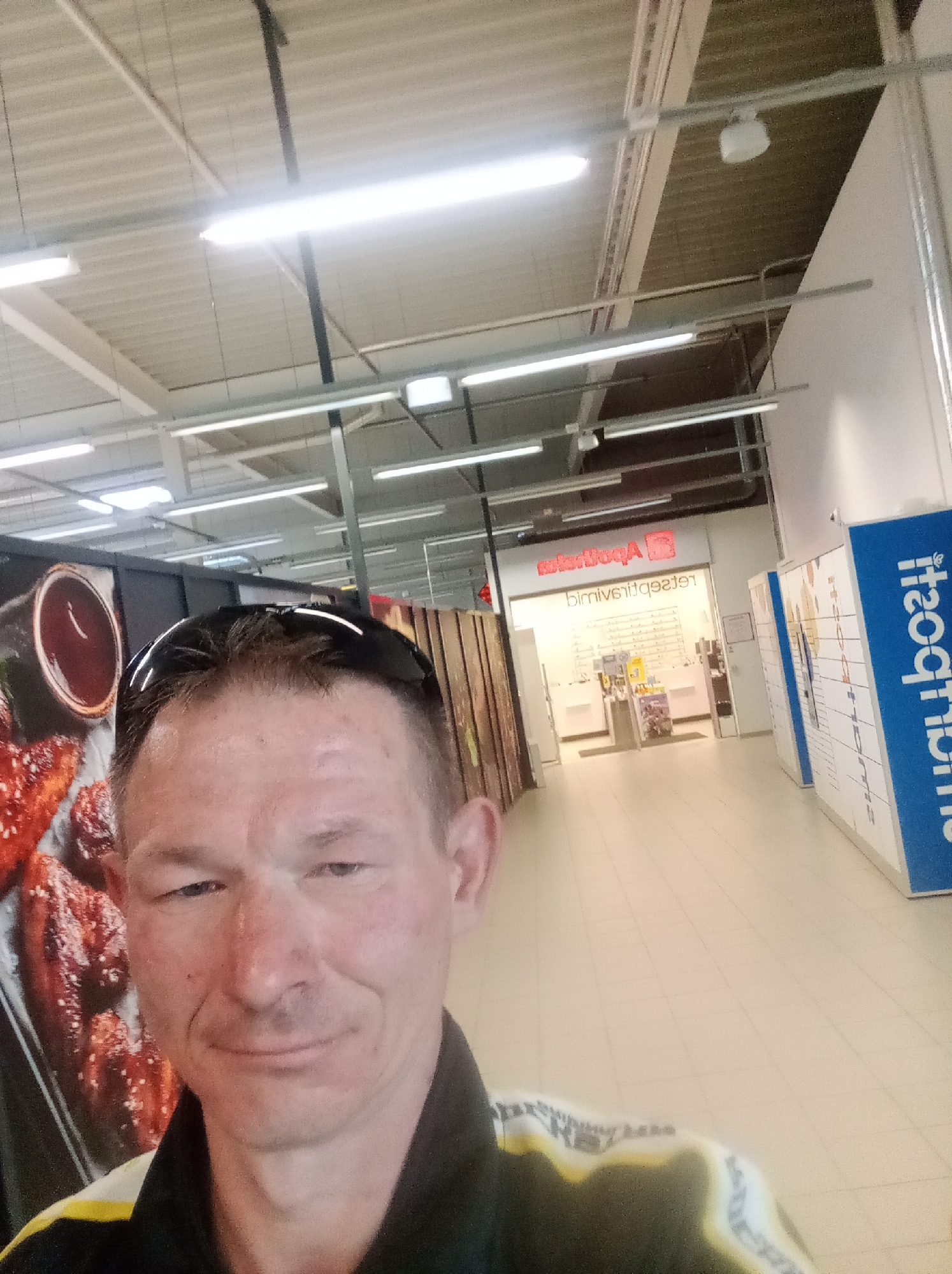 A man is taking a selfie in front of an "Apotheka" store in Tapa, Estonia. The man is wearing sunglasses on his head and a black and yellow jacket. The store has a large window displaying shelves of products. To the right of the store are blue and white lockers. The flooring is tiled.