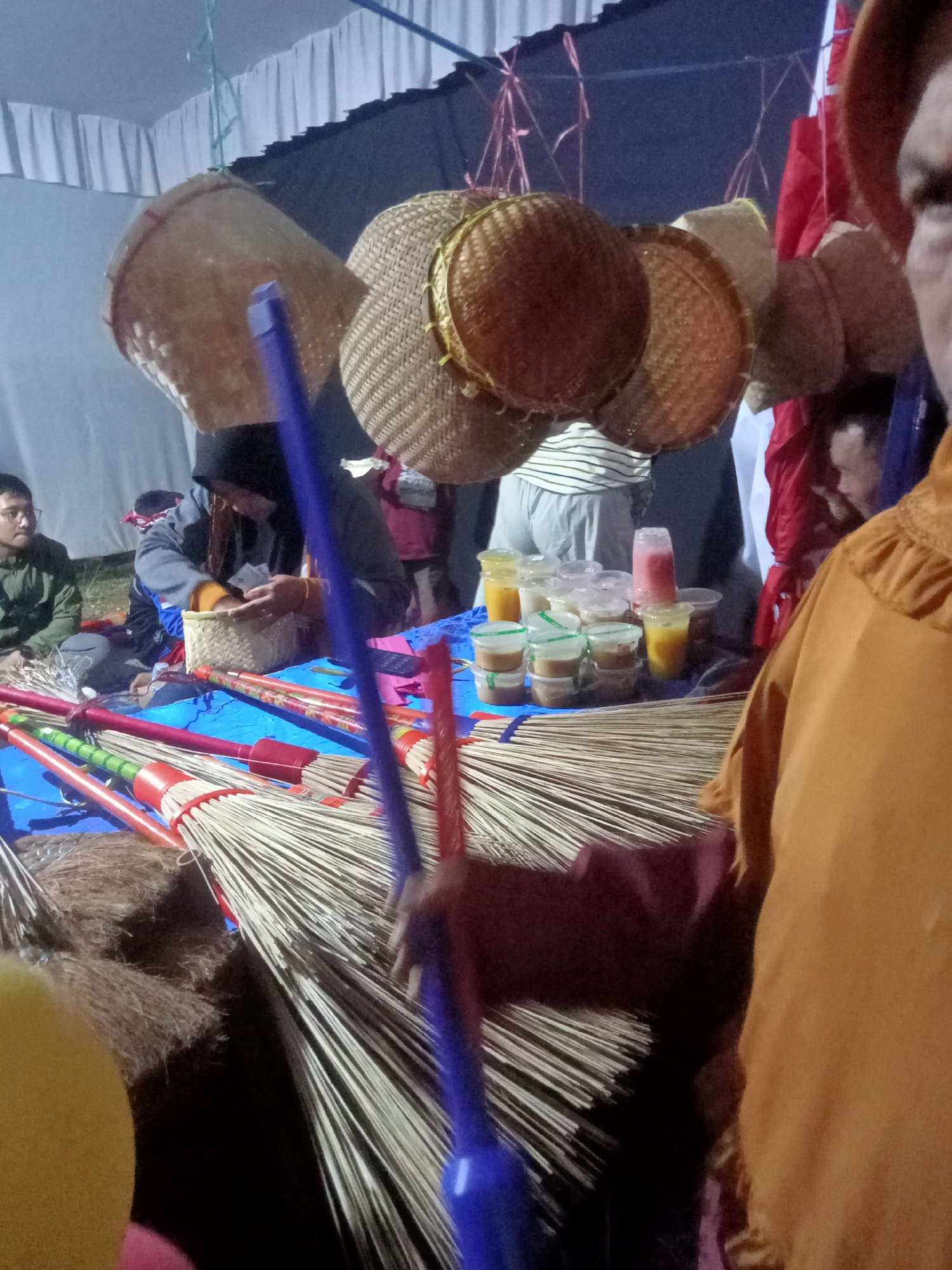 In Pule, Indonesia, a market stall displays numerous brooms with wooden handles in red and green, neatly arranged on a blue tarp. Several small plastic cups containing colorful liquids, likely beverages or desserts, are placed behind the brooms. Above the stall, woven baskets hang suspended, creating a rustic canopy. In the background, people are gathered, with one individual wearing a dark headscarf and another a green jacket. A person in an orange garment is prominently positioned in the foreground, their face partially obscured. The scene appears to be an outdoor market or fair.