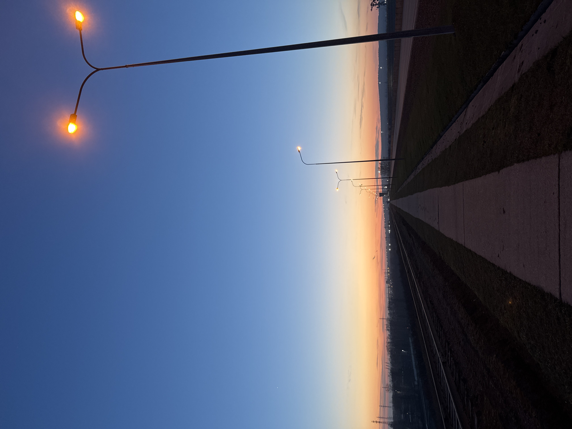 This image, presented vertically, captures a tranquil twilight scene, likely at dusk, in Chaikovsky, Russia. The vast sky dominates the left portion of the frame, displaying a clear gradient from a deep, dark blue at the top to a warm, soft orange and yellow along the horizon line, which is visible towards the bottom right of the image.
In the upper left foreground, a prominent street light with two illuminated, warm orange bulbs extends from a dark pole. This pole stretches diagonally upwards across the blue sky. Further into the distance, a series of other street lights or utility poles, each emitting a softer white glow, line a path or road that recedes towards the horizon.
On the right side of the image, a straight, lighter-toned path or road runs parallel to a strip of darker ground, possibly grass or unpaved land. Adjacent to these, on the far right, are what appear to be railroad tracks, their parallel lines converging into the distance. The distant horizon, bathed in the orange glow of the setting sun, shows faint outlines of what might be industrial structures or distant landscape features.
The scene is static and devoid of any human presence or explicit activity, creating a sense of solitude and peace. The weather appears clear, with no clouds visible across the expansive sky. No discernible text is present in the image.