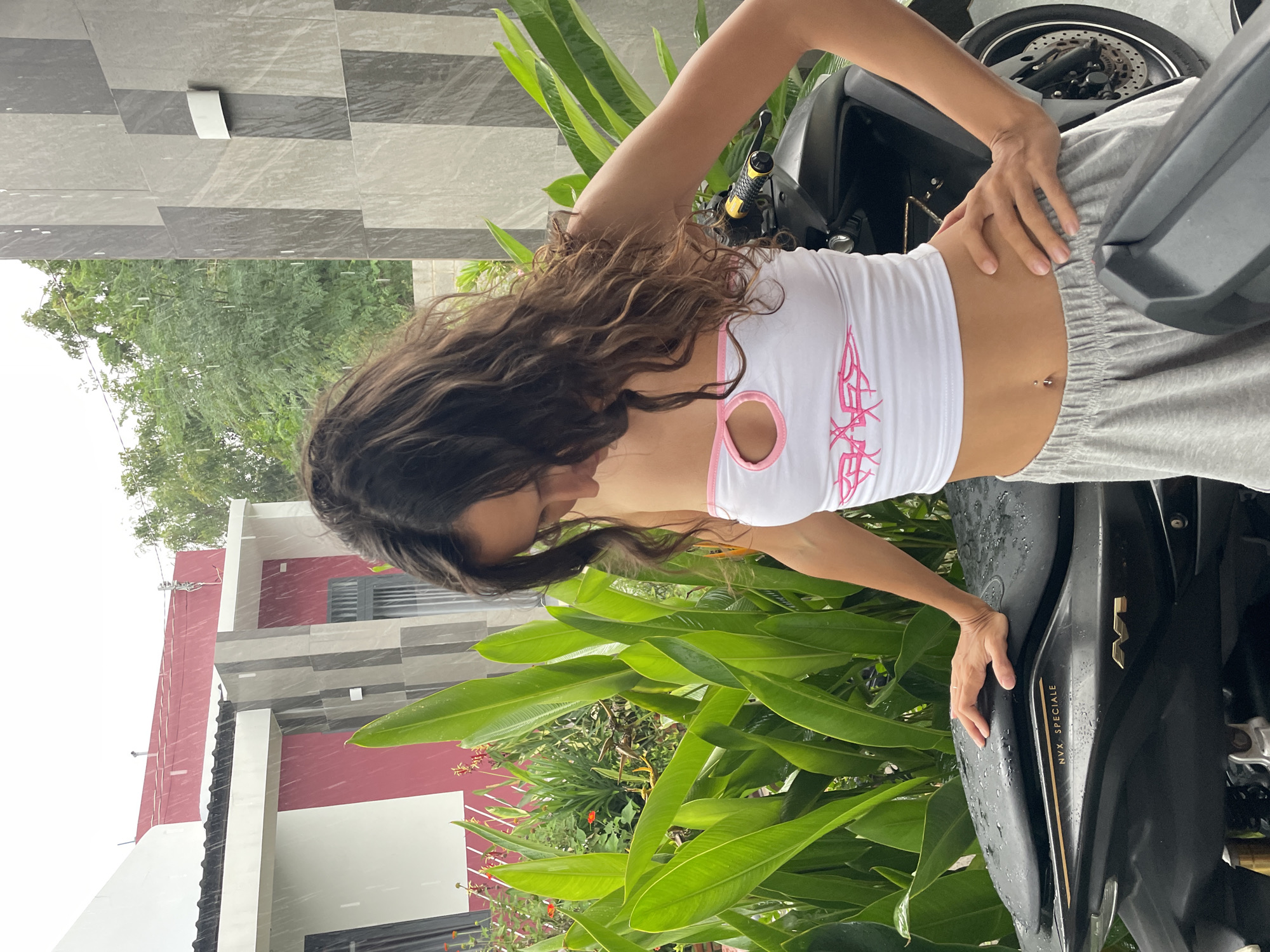A young woman with long, dark, curly hair, wearing a white tube top with pink trim and a pink graphic design, and gray sweatshorts, stands with her hands on her hips and the other resting on the seat of a black motorcycle. The motorcycle has "NVX SPECIALE" printed on its side. The woman has a belly button piercing. It is raining, with visible raindrops falling. The background features lush green foliage and a modern building with white and red sections. The scene appears to be in Mũi Né, Vietnam, as indicated by the architecture and tropical vegetation.