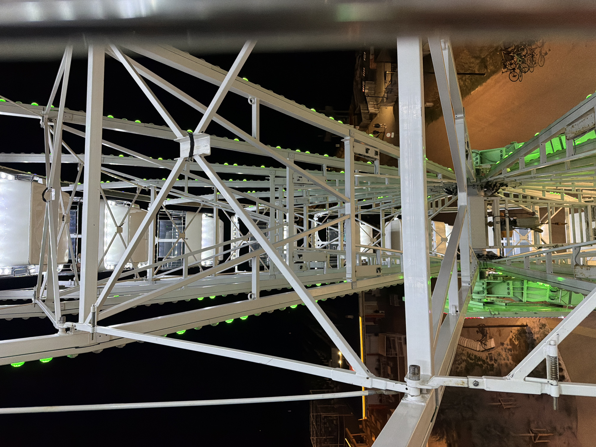 This is a close-up, low-angle, nighttime photograph of the white metal structure of a Ferris wheel in Gatteo, Italy. The Ferris wheel is illuminated with green lights that line its outer circumference and some of its internal structure. The structure is complex, with numerous beams and supports visible. The black night sky serves as the backdrop for the illuminated wheel. Below the Ferris wheel, a dirt path is visible, with a cluster of bicycles parked on the right side of the path. Two figures, appearing to be adults, are walking away from the camera on the path. In the background, there are some low-lying structures and fencing, suggesting an amusement park or recreational area. The overall impression is of an active amusement park at night.
