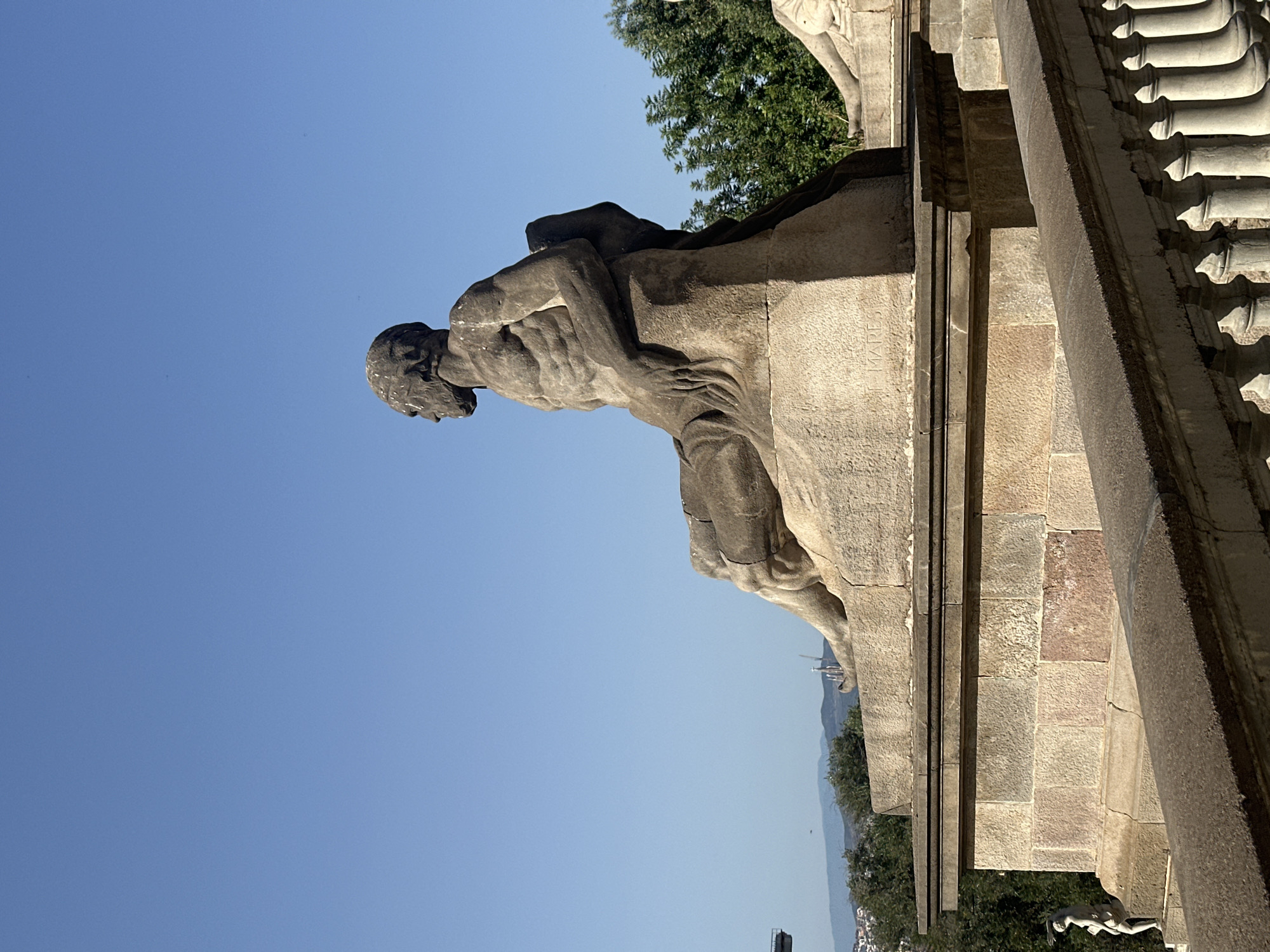 This media file depicts a stone sculpture of a man in a pensive or distressed pose, set against a clear blue sky. The sculpture is detailed, showing muscular definition and a thoughtful expression on the man's face. He appears to be leaning or struggling, with his arms wrapped around himself or supporting his head. The sculpture is positioned on a stone structure that suggests a monumental setting, possibly a monument or a public building.
In the background, there are glimpses of lush green trees and a distant landscape that includes some structures, hinting at an urban environment. The lighting suggests it is daytime, with bright sunlight casting shadows on the sculpture and the stone architecture. The overall mood conveyed by the sculpture is one of introspection or suffering.
Visible text on the stone structure reads "MARES". The image is oriented vertically, with the sky occupying the upper left portion and the sculpture and architecture filling the rest of the frame. The setting appears to be outdoors, likely in a historical or culturally significant location. Given the location context of Barcelona, this could be a part of a larger monument or a significant architectural feature within the city.