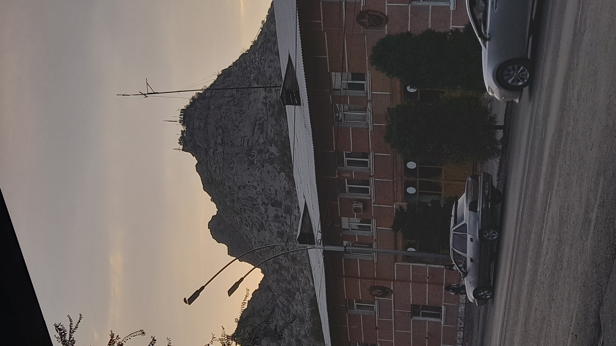 A medium shot captured from a low angle shows a mountain range behind a building in the city of Unknown. The mountain range is covered in large, gray rocks and a transmission tower is visible on the peak. The building is a brick structure with a white roof and numerous windows. Trees are planted in front of the building. Two cars are parked on the street in front of the building, and a person is walking in the distance. The sky is overcast and the sun is setting.