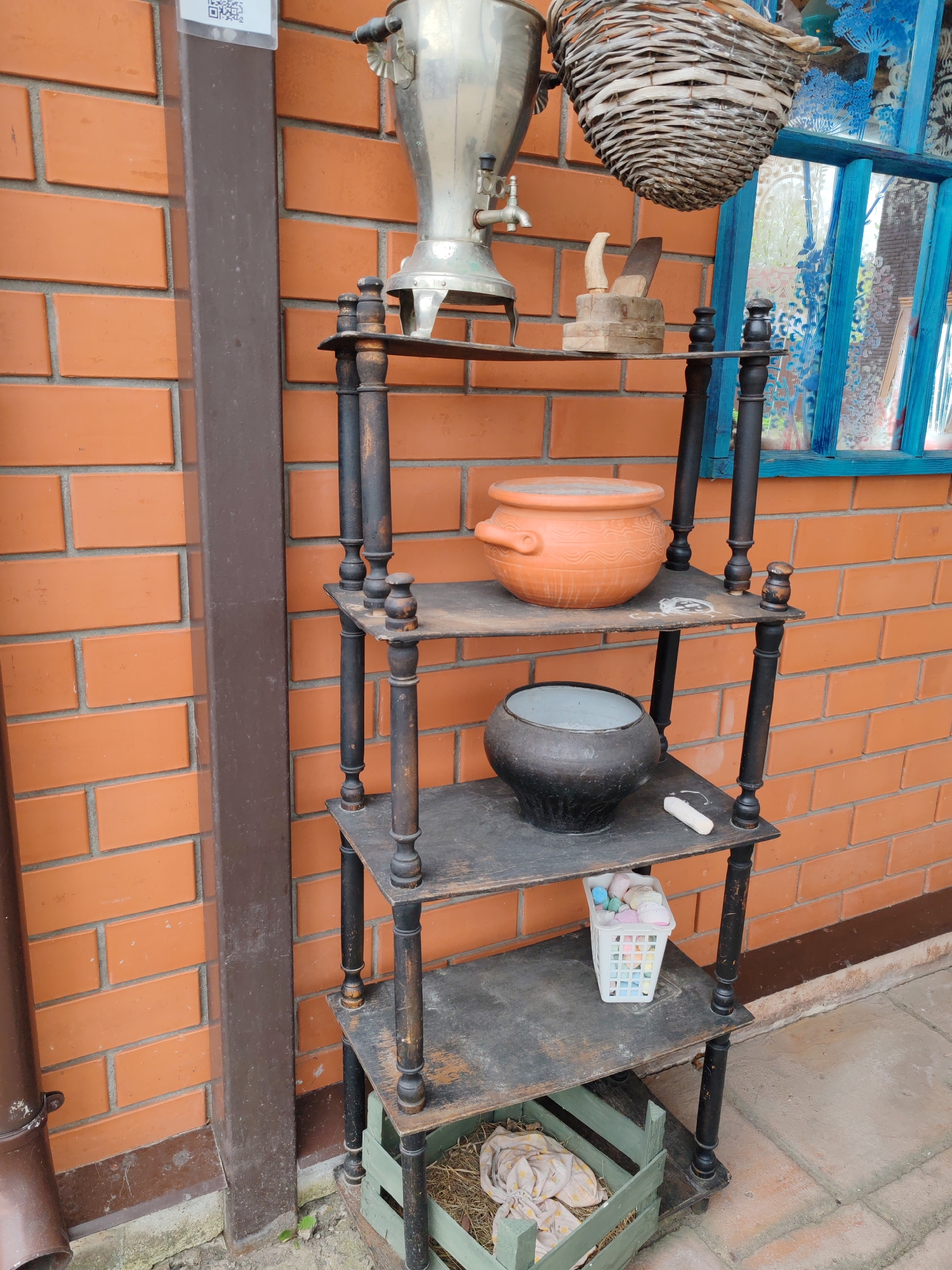 A four-tier wooden shelf sits against a brick wall in Tver, Russia. The shelf is dark, aged, and shows signs of wear.
From top to bottom, the shelf displays: a silver samovar, a wooden chopping block with a knife, a woven basket hanging overhead; an orange ceramic pot; a black metal pot with a light gray interior, a piece of white chalk; a small white plastic basket containing marshmallows; and a green wooden crate filled with straw and soft white fabric.
To the left of the shelf is a brown drainpipe attached to the brick wall. Above the shelf is a window with a blue frame. The ground is a mix of concrete and dirt.