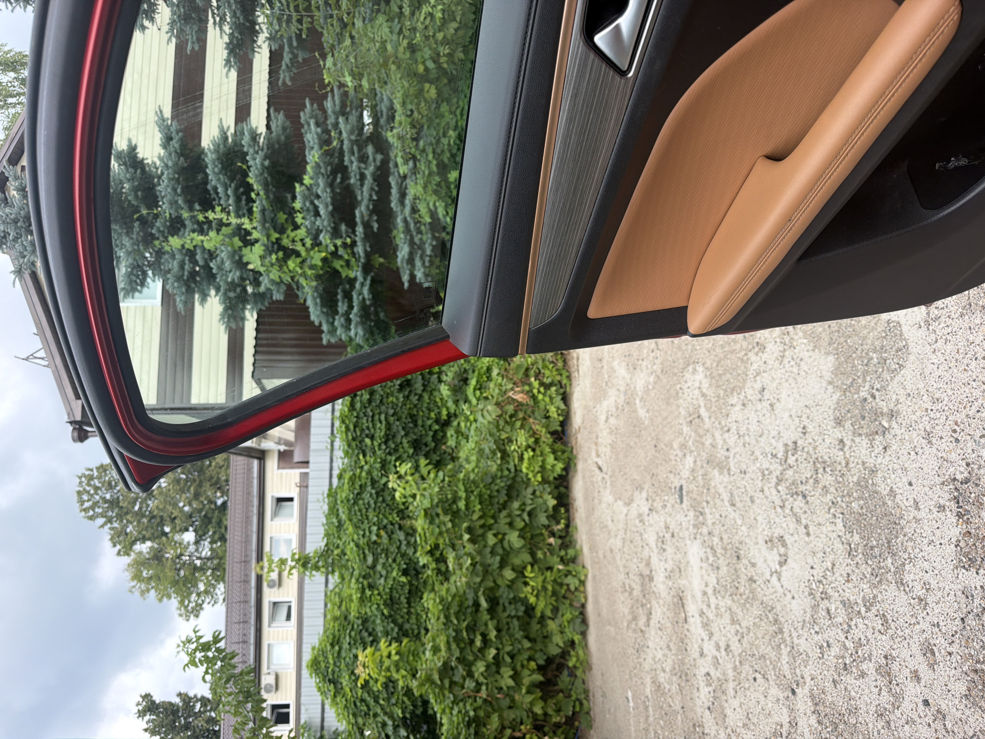 The image shows the interior of a car door, with the door open, revealing an outdoor scene. The car's interior features a tan leather armrest and a metallic trim accent. Outside, a patch of concrete or gravel is visible in the foreground, leading to a lush green hedge and trees. In the background, a building with light-colored siding and multiple windows can be seen. The sky is overcast. The image does not provide any specific details to indicate its location in Kazan, Russia.