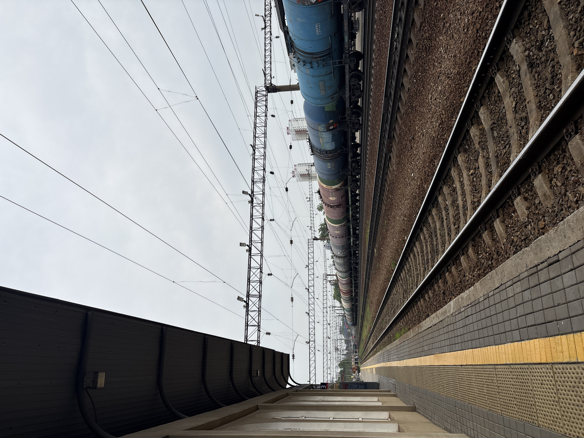 The image depicts a railway station platform, likely in Khabarovsk, Russia, given the style of railway cars and infrastructure. A long train consisting primarily of tank cars stretches along the tracks. Overhead power lines supported by metal pylons run parallel to the train.
The platform has a textured surface and a yellow safety line. A set of steps leads up to the platform from a lower level. A dark, curved sound barrier wall is adjacent to the steps. The sky is overcast. There are no people visible in the immediate foreground.