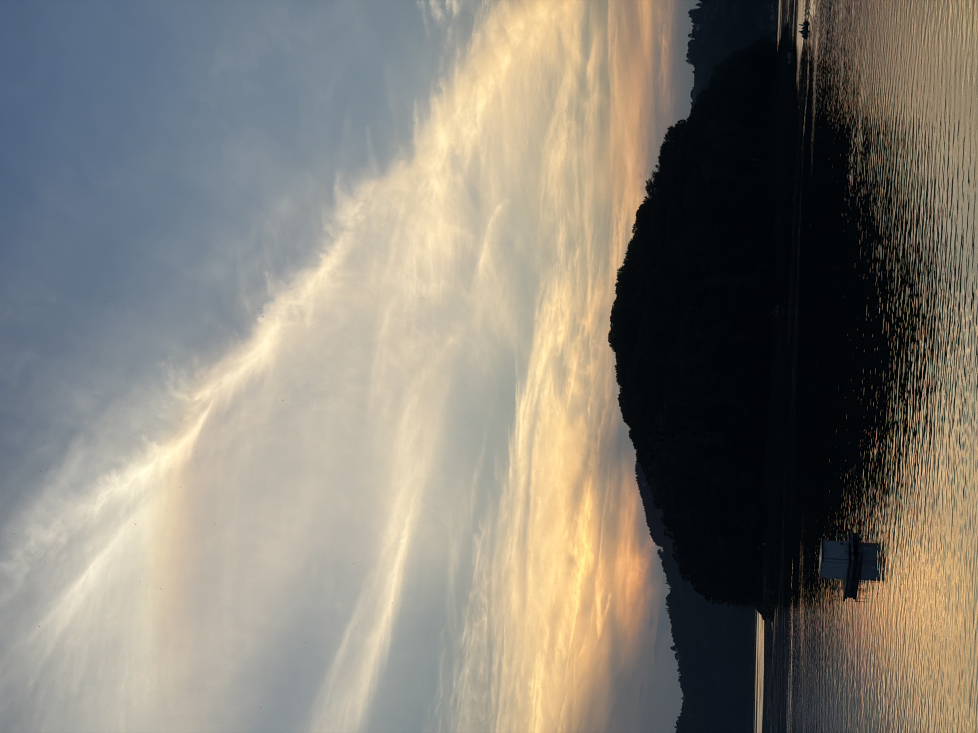 A serene sunset over Lake Rożnowskie in Gródek nad Dunajcem, Poland, captures a tranquil scene. The sky is painted with a gradient of soft orange and yellow hues near the horizon, transitioning to a pale blue at the top, streaked with delicate, wispy clouds. In the foreground, the still waters of the lake reflect the warm colors of the sunset, creating shimmering patterns. A small, white floating structure, possibly a buoy or a small hut, sits on the water to the left, its reflection clearly visible beneath it. Silhouetted against the vibrant sky, a large, tree-covered island dominates the middle ground, its dark outline sharp and defined. In the distance on the right, a small boat with two occupants can be seen navigating the water. The surrounding hills on the far bank are also silhouetted in dark tones, creating a dramatic backdrop to the illuminated sky and water. The overall impression is one of peace and natural beauty.