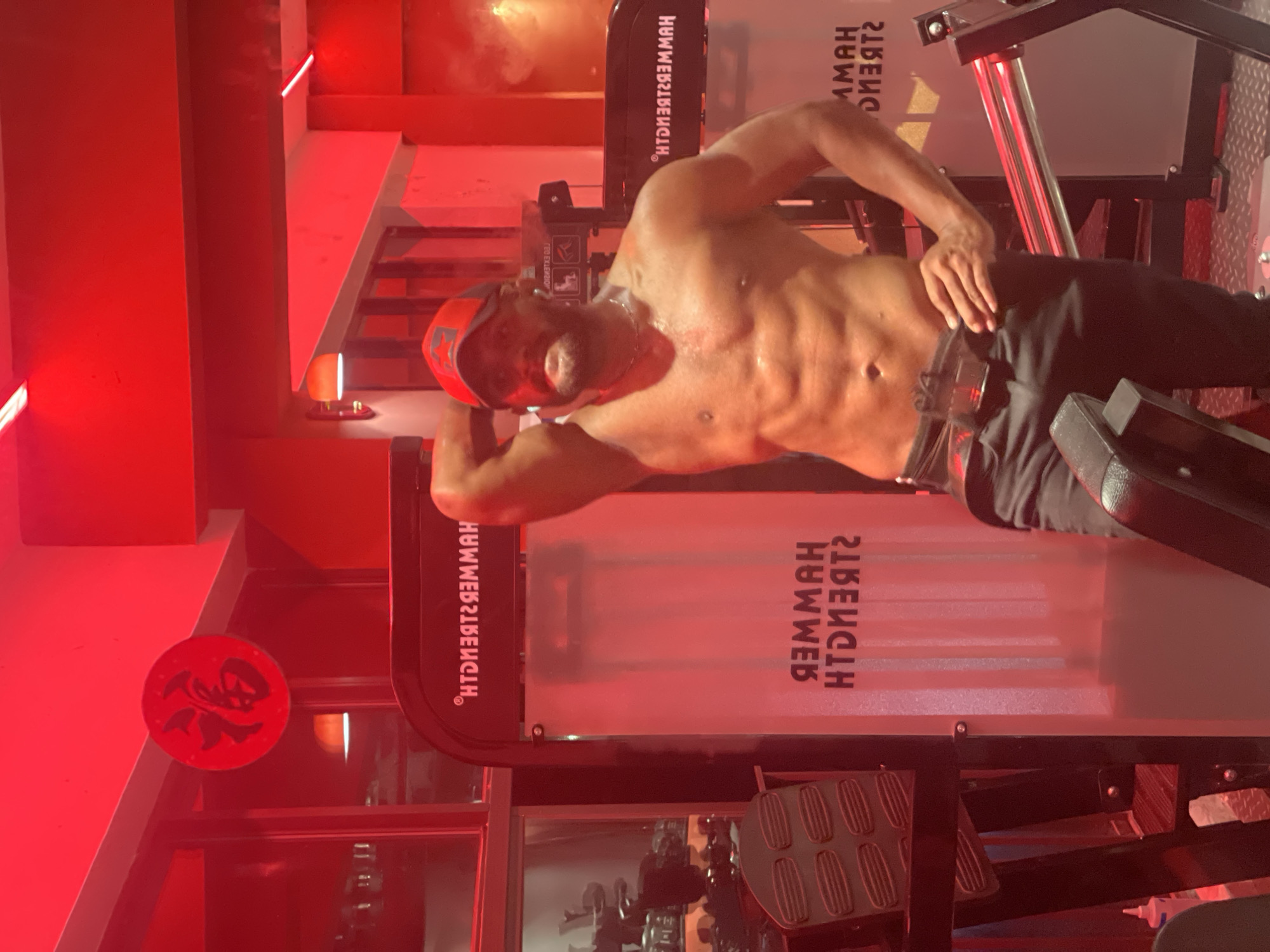 A man is posing shirtless in a gym with red lighting. He is wearing a red baseball cap and black pants. The gym appears to be a Hammer Strength gym, as indicated by the branding on the equipment. There is a circular red decoration with Chinese characters on the wall in the background. The lighting casts a strong red hue over the entire scene. The man is standing in front of weight machines.