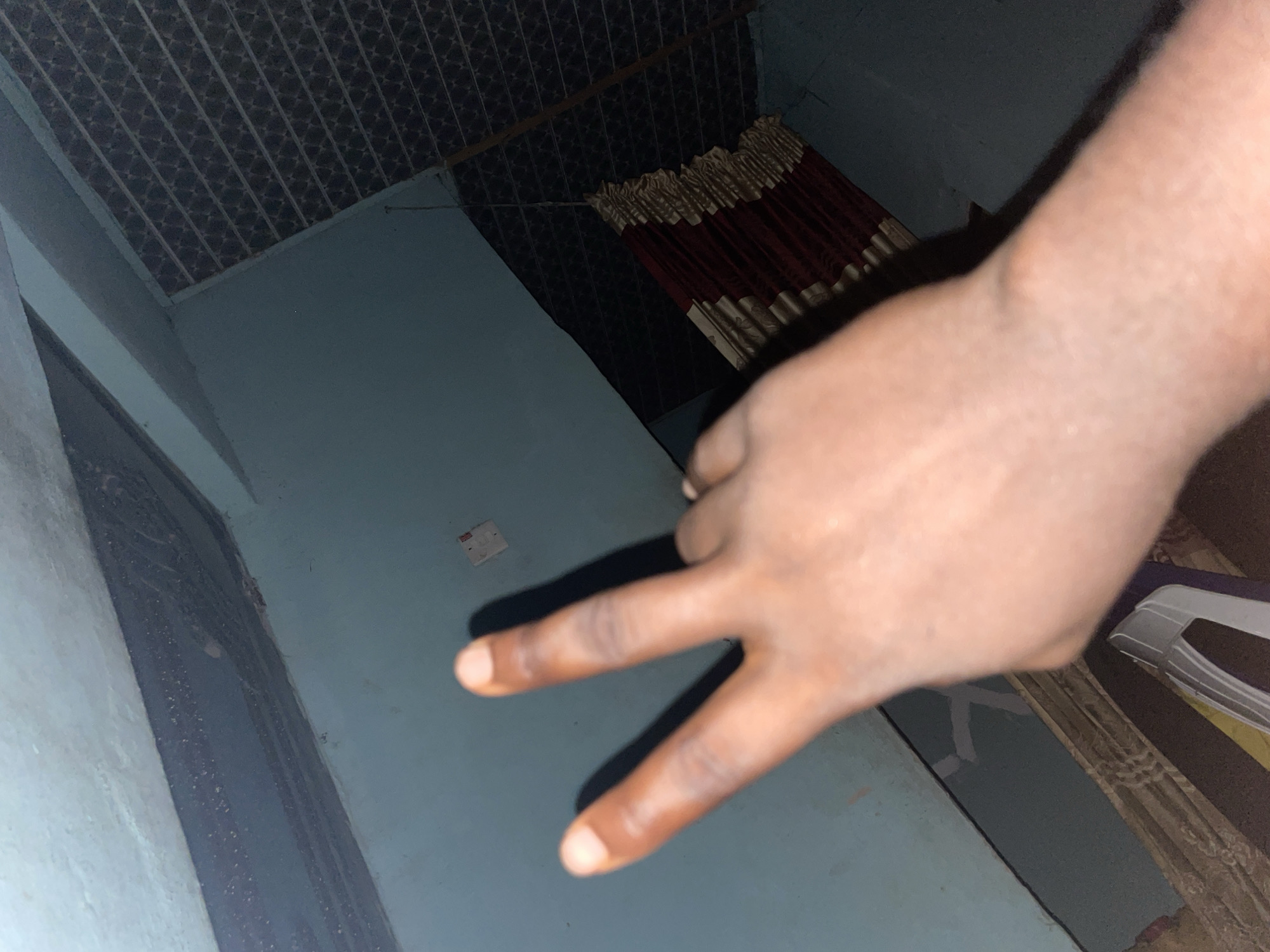 A person's hand is shown, with two fingers extended in a peace sign. The hand is in the foreground, and the background appears to be an interior space. The walls are painted a light blue. There is a dark, textured ceiling with what looks like a suspended fixture covered by red and gold fabric. In the background, a white object, possibly part of a piece of furniture, is visible. The lighting suggests it may be nighttime or a dimly lit room.