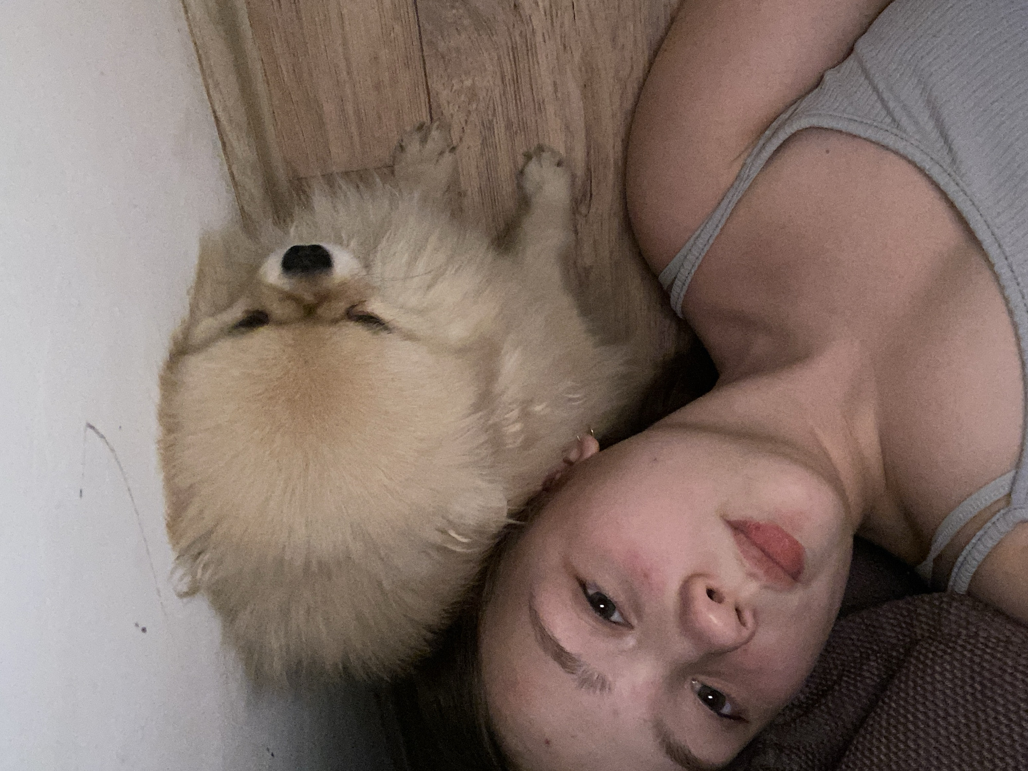 A light-skinned person with light brown hair is lying on a brown fabric surface, with a light brown Pomeranian dog lying next to the person on a wooden floor. The person is wearing a grey tank top. The room is indoors, with a white wall to the left and a wooden floor in the background. The location is unknown.