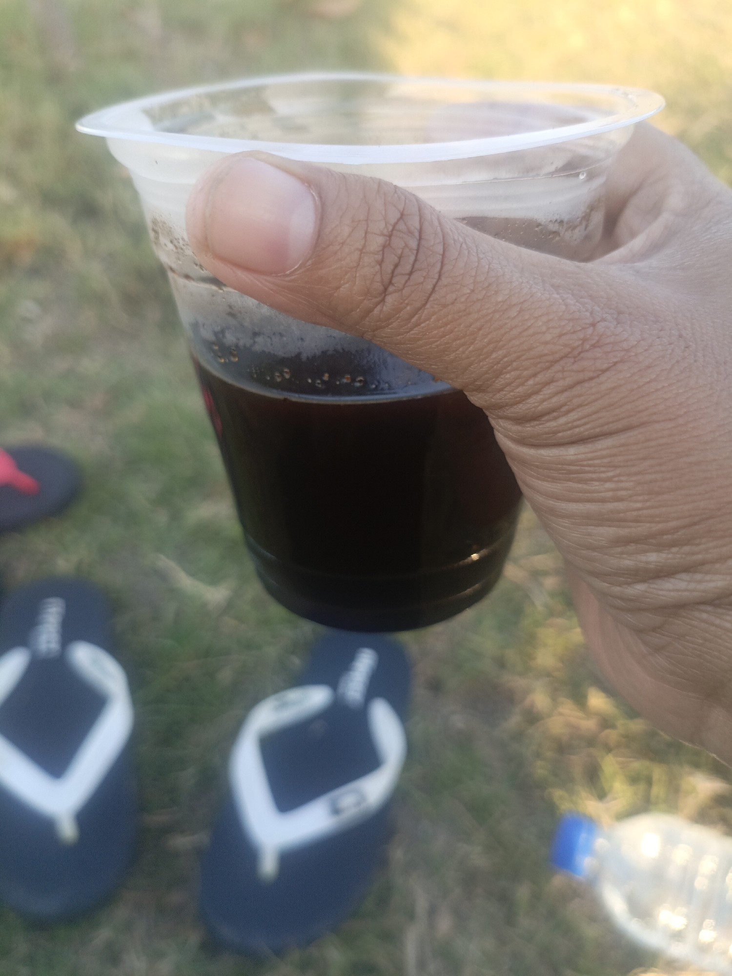 A hand is holding a clear plastic cup filled with a dark liquid, possibly coffee or soda. The cup has a white plastic lid. The background is blurry and appears to be outdoors, with green grass and some yellowed vegetation visible. In the lower left of the frame, two dark flip-flops with white straps are on the ground. To the right, a blurry plastic water bottle with a blue cap is also visible. The scene suggests a casual outdoor setting. No specific location in Lewoleba, Indonesia, can be identified from the image.