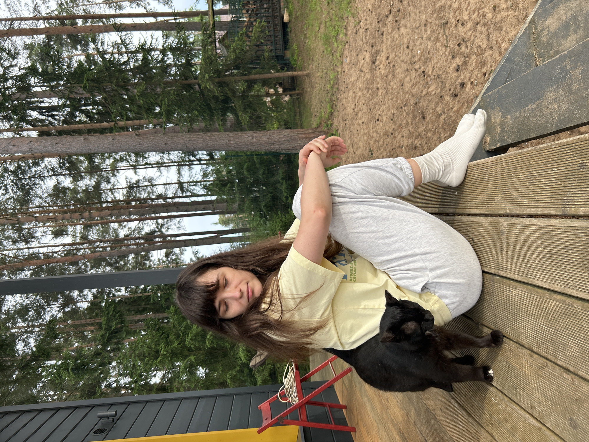 In this media file, a young woman with long brown hair and bangs, wearing a light yellow t-shirt and grey sweatpants with white socks, is relaxing on a wooden deck. She is curled up, with her eyes closed and her head resting on her arm, appearing to be asleep or deep in thought. A black cat is nestled beside her, also seemingly at rest. The scene is set outdoors, amidst a backdrop of tall pine trees, suggesting a natural, possibly wooded, location. The wooden deck, some steps, and a red foldable chair are visible, hinting at a casual outdoor living space, possibly a porch or patio of a house or cabin. The ground beyond the deck is covered with sandy soil and small pebbles, further reinforcing the natural setting. The lighting suggests it is daytime, with soft natural light filtering through the trees. The overall mood is one of tranquility and peacefulness. There is no visible text in the image. The location context is Shepelyovo, Russia.
