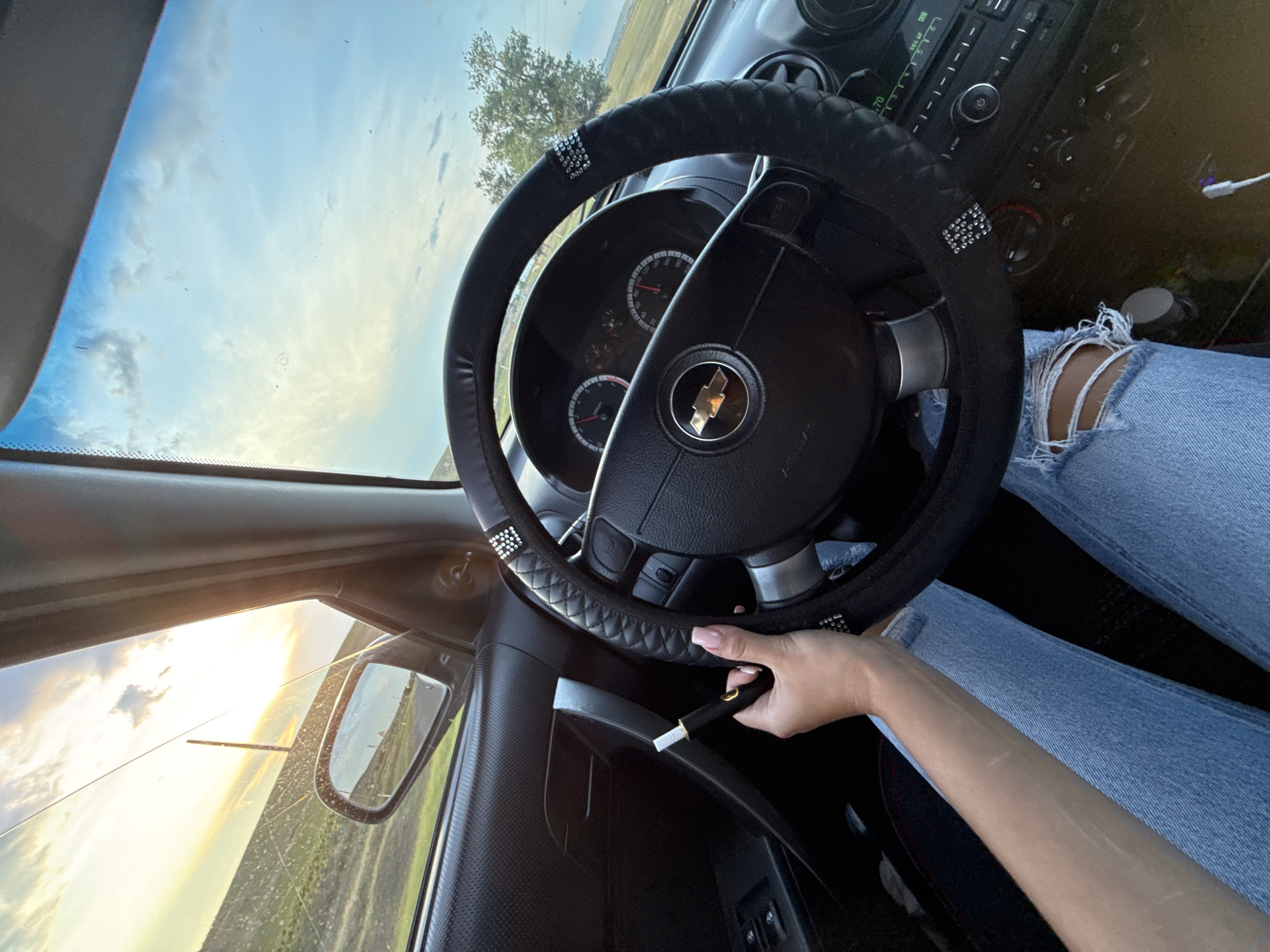 A POV shot from the interior of a car shows a person's left hand holding a dark electronic cigarette device with a white tip near the steering wheel. The person is wearing light blue ripped jeans and is seated in the driver's seat. The car's steering wheel is black with a textured grip and is adorned with rhinestones. The dashboard has a Chevrolet emblem in the center of the steering wheel and displays a speedometer and tachometer. The car's window offers a view of a sunset with clouds in a blue sky, and a rural landscape with fields and a distant tree. The image is taken during golden hour. The car appears to be located on a road in the vicinity of Iordanovca, Moldova, based on the provided context.