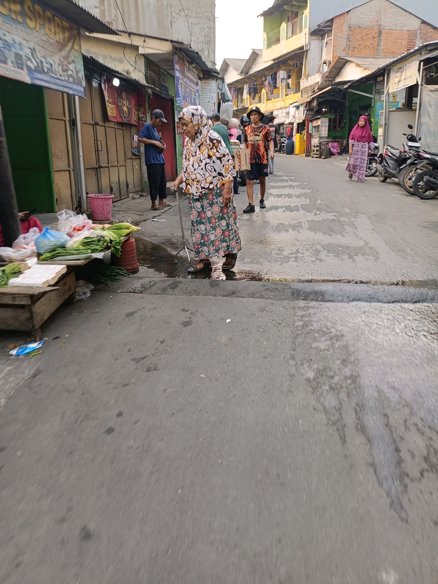 The image depicts a street scene in Tangerang, Indonesia, likely a market area. In the foreground, an elderly woman wearing a headscarf and a floral patterned skirt is walking with the aid of a cane. To her left, a wooden stall displays various fresh vegetables like long beans, corn, and tomatoes. Further down the street, several individuals are visible, including a man in a t-shirt and shorts, and a woman in a purple headscarf and dress. Motorcycles are parked along the side of the street. The buildings lining the street appear to be residential or small commercial establishments, with multiple stories and balconies. The wet asphalt surface suggests recent rain or ongoing water activity. The overall atmosphere is one of a bustling, everyday urban environment.