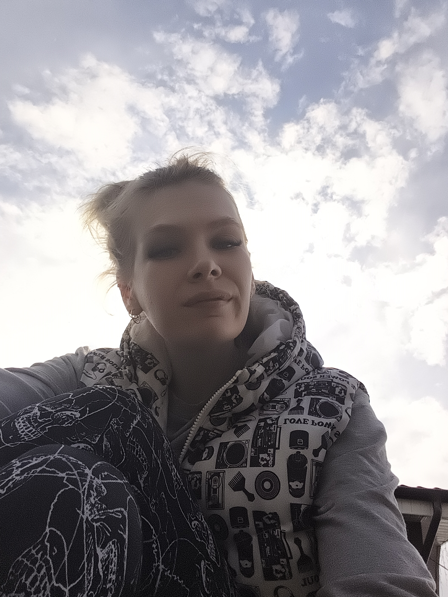 A low-angle, medium shot captures a young woman posing outdoors. She has fair skin, light hair pulled back, and wears dark eye makeup. She is dressed in a hooded vest with black and white prints over a gray long-sleeved shirt. Her black pants have a white, skeletal pattern. The sky, filled with white clouds against a blue backdrop, dominates the upper half of the frame. A portion of a building roof is visible in the lower right corner. The photo's location is specified as Русский Вожой, Russia.