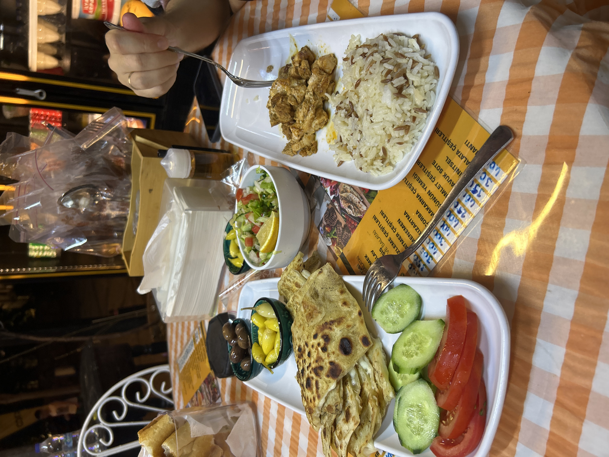 A high-angle medium shot captures a dining table at an outdoor restaurant in Ortaca, Turkey, with an orange and white checkered tablecloth. The table is laden with Turkish cuisine. In the foreground, a white rectangular plate holds a stack of folded flatbread, possibly gözleme, with visible cheese filling, accompanied by sliced cucumbers and tomatoes. To the right, another white rectangular plate contains a portion of seasoned, diced chicken and a mound of mixed rice. A fork is lodged in the chicken. In the background, several small bowls hold olives, pickled peppers, and a salad with lemon. A stack of napkins, a bottle of oil, and a cell phone rest on the table. A partially visible person's hand with a ring is seen reaching for the food with a fork. The restaurant's interior, with illuminated shelves and wrapped food items, is visible in the blurred background.