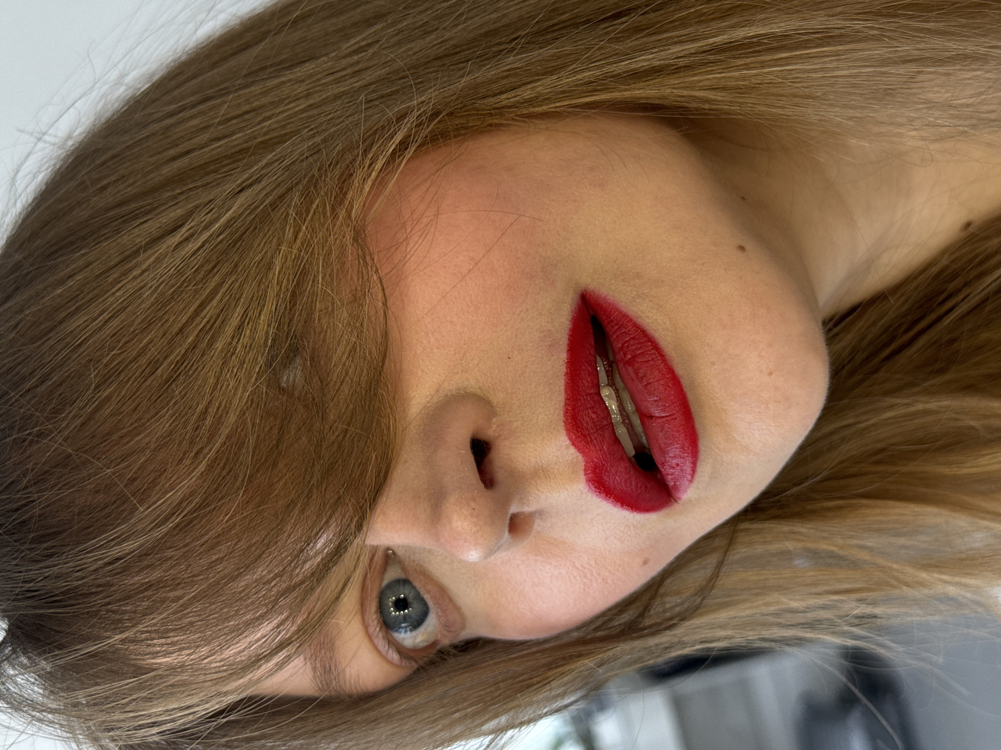 The image is a close-up of a woman's face, positioned at an oblique angle. Her hair is light brown and partially covers her forehead. She has one visible eye, which is blue. Her lips are painted a vibrant red. The background is a plain, light color. There is no information in the image that would indicate the location is Izhevsk, Russia. The image solely focuses on the woman's facial features.