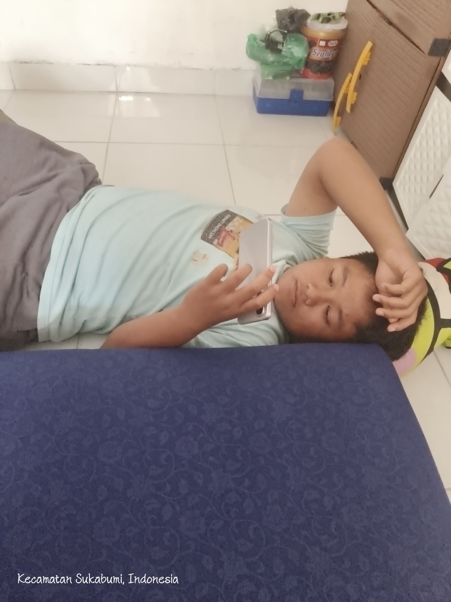 A young boy is lying on the floor, propped up on his left elbow, looking at his phone. He is wearing a light blue t-shirt and grey shorts. The floor is tiled in a light color. In the background, there is a stack of items, including a blue and clear plastic box, a yellow object, and a brown cardboard box. To the right of the boy, part of a colorful object, possibly a pillow or cushion, is visible. A blue textured surface fills the bottom of the frame. Text at the bottom of the image reads "Kecamatan Sukabumi, Indonesia."