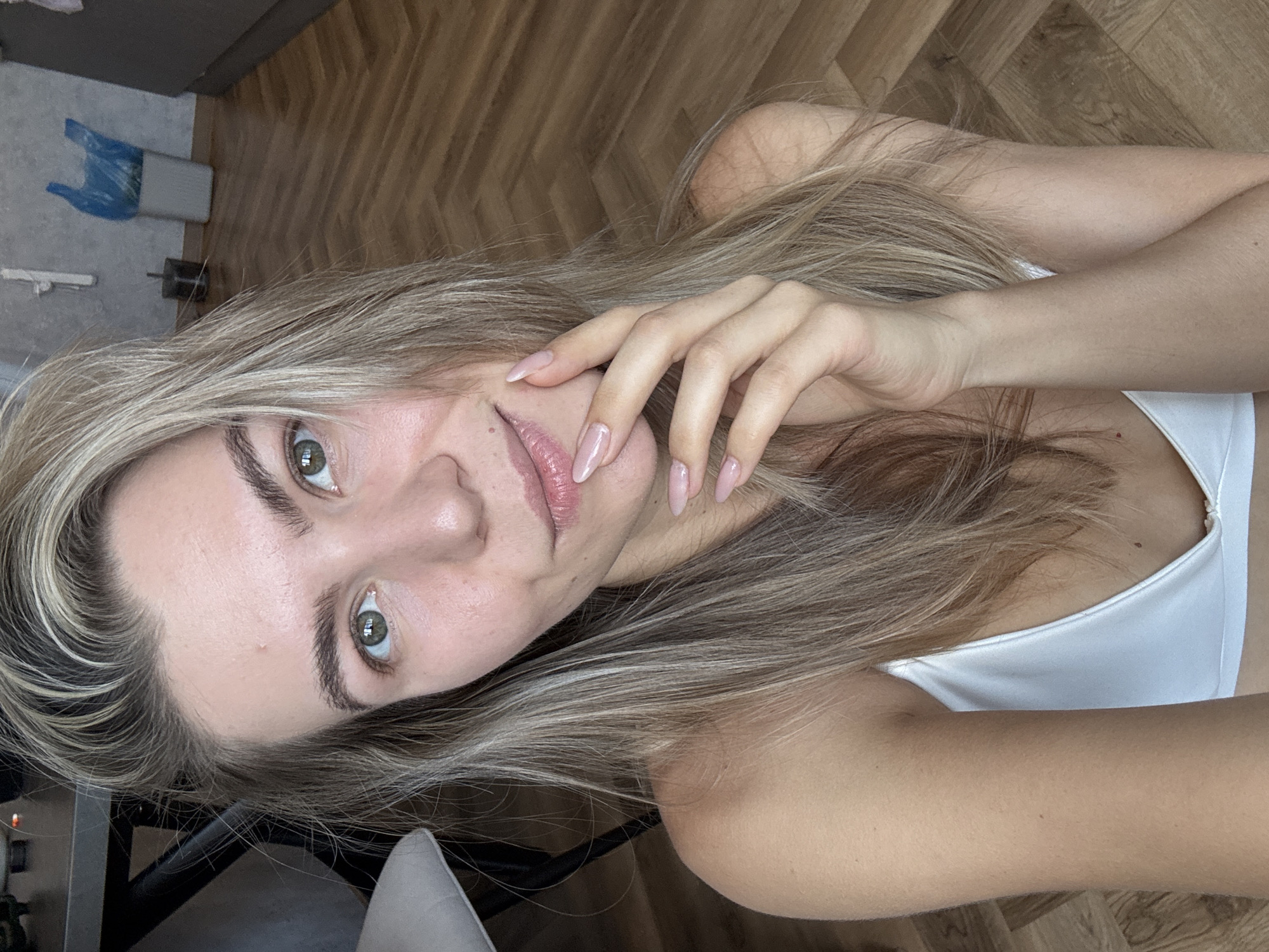 A fair-skinned woman with shoulder-length blonde hair and green eyes is posing for a selfie. She is wearing a white top. Her right hand is touching her face. The background consists of a wooden floor and glimpses of other objects like a chair and what appears to be a cleaning supply in the corner. The location is a building in the city of Unknown, Unknown.