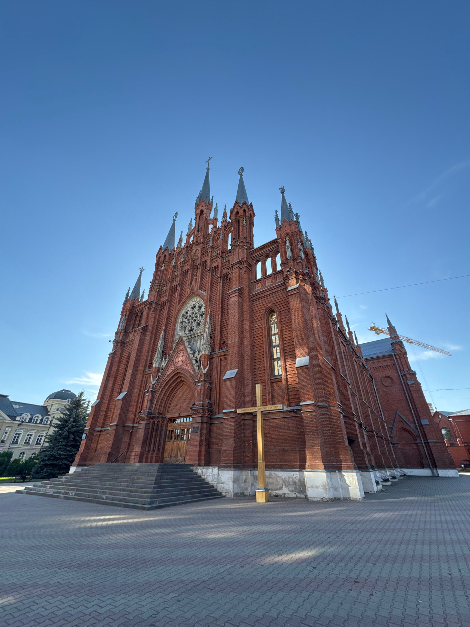 The image shows the Cathedral of the Immaculate Conception in Moscow, Russia, a large red brick Gothic Revival Catholic cathedral located at 12 Malaya Gruzinskaya Street. The sun is shining, casting shadows on the building and the paved ground in front of it. A wooden cross stands prominently in the foreground, inscribed with Russian text. The cathedral's facade features a large rose window above the entrance, flanked by two tall spires. To the left of the cathedral, a portion of another building and a tall evergreen tree are visible. In the background, a construction crane is present, indicating ongoing development in the area. The sky is clear and blue.