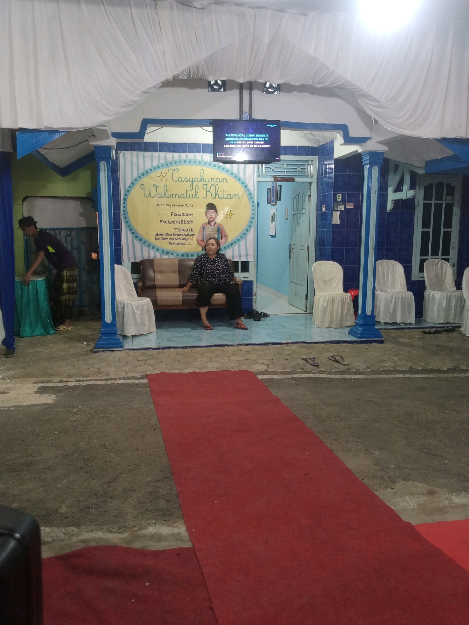 The image depicts an outdoor event space, possibly a front yard or entrance to a building, in Rejasari, Indonesia. A red carpet leads from the foreground into the scene, where a covered structure is set up. This structure appears to be decorated with blue and white striped fabric.
On the left side of the structure, a man wearing a hat and traditional Indonesian pants is seen arranging a teal-colored cloth on a table. To the right of him, inside the structure, a woman is seated on a brown and beige couch. She is wearing a dark patterned top and black pants, and her feet are bare.
Behind the woman and on the couch, a banner with yellow and blue accents is displayed. It features text in Indonesian, including "Tasyakuran Walimatul Khitan" and names that appear to be "Fauzan" and "Fatahillah". A picture of a young boy is also visible on the banner.
In front of the structure, several white chairs covered with white fabric are arranged. To the right of the entrance, a blue tiled wall is visible, with a door and a window. A "9" is displayed on the wall.
Above the structure, a large white draped fabric is suspended. A flat-screen television is mounted on the wall, displaying text and images. The ground in front of the structure is paved with concrete and appears to be somewhat rough. Scattered footwear, including flip-flops, is visible on the ground. The overall impression is of a celebration or gathering taking place.