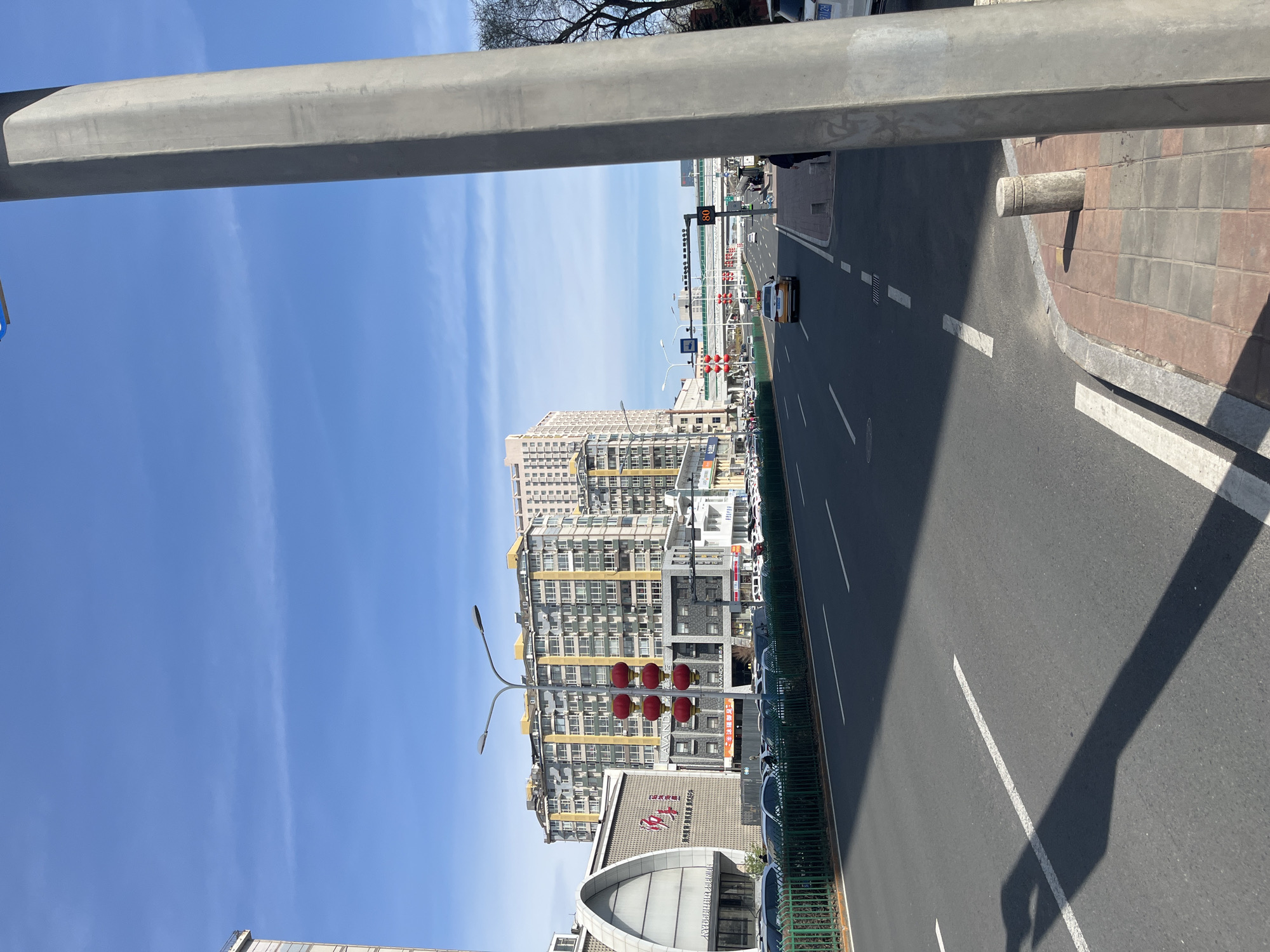 The image depicts a street scene in Changchun City, China. The perspective is taken from an elevated position, possibly from a pedestrian bridge or overpass. The view is oriented along a multi-lane road with a concrete median separating the lanes. The road surface is asphalt with painted white lane markings. Tall buildings with a yellow and white color scheme line the left side of the road, interspersed with lampposts and a traffic light with red globes. The sky is clear blue with some light clouds. A grey horizontal structure, likely part of the overpass, occupies the upper portion of the frame. The edge of the sidewalk is visible to the right with a brick curb and a metal barrier lining the road. A few vehicles are visible on the road, including a yellow car.