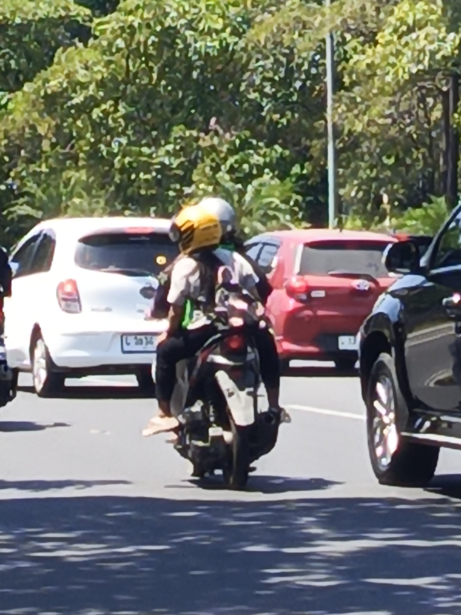 The image shows a street scene with several vehicles and people. In the foreground, a motorcycle with two occupants is stopped, with the rear wheel and part of the frame visible. The passenger is wearing a yellow helmet and a light-colored shirt with green accents, and the driver is wearing a dark helmet. Behind the motorcycle, a white car is parked or stopped, with its rear visible. Further in the background, a red hatchback is visible, and to the right, the side of a dark SUV is present. The environment appears to be outdoors, with lush green foliage forming a backdrop, suggesting a location with abundant vegetation. The lighting indicates it is daytime, with shadows cast on the road. The license plate on the white car is partially visible and appears to have Thai or similar characters. The overall scene depicts a moment of traffic or a pause in movement on a city street. Based on the style of vehicles and potential license plate characters, the scene is likely in Southeast Asia. It has been identified that this image was taken in Surabaya, Indonesia.