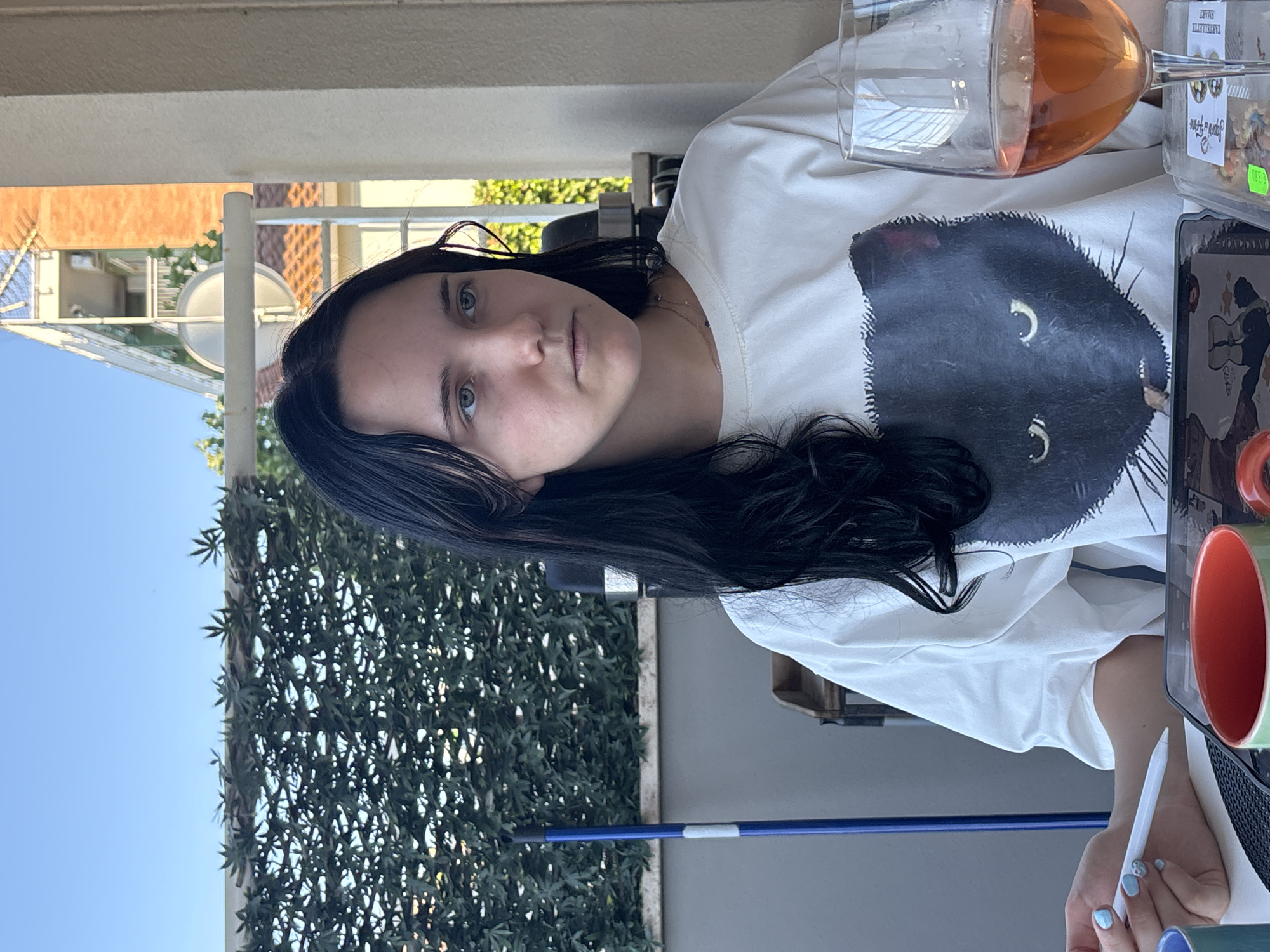 A young woman with long, dark hair and light blue eyes is seated outdoors, likely on a balcony or patio in Cupra Marittima, Italy, as indicated by the architectural style of the buildings visible in the background. She is wearing a white t-shirt with a graphic of a black cat on it. To her right, a glass of rosé wine is placed on the table. Her left hand rests on the table, holding a stylus, and she appears to be interacting with a tablet in front of her. In the background, a green hedge provides a backdrop, and a blue pole is visible to the left. The environment suggests a bright, sunny day.
