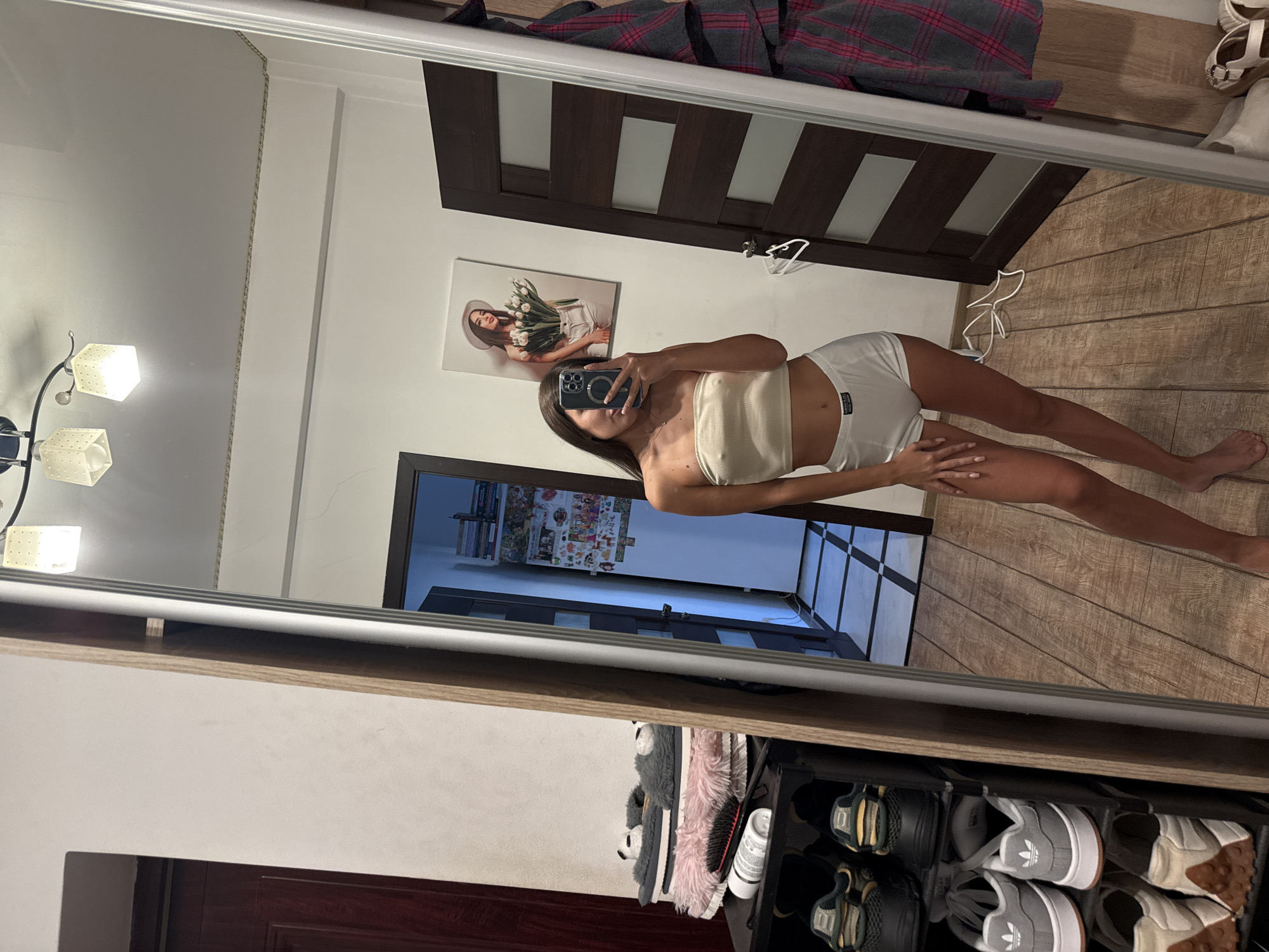 A young woman, with long dark hair, is standing in front of a full-length mirror in what appears to be a bedroom or dressing area. She is wearing a beige bandeau top and white boxer shorts. She is holding a smartphone in her right hand, taking a selfie.
The room has light-colored walls and a wooden floor. To the left of the mirror, there is a shoe rack filled with various shoes and some fuzzy slippers. To the right of the woman, a dark wooden door is visible, with a piece of artwork hanging on the wall above it. The artwork depicts a woman holding a bouquet of flowers. A plaid garment is hanging on the right side of the mirror.
The overall impression is that of a private living space, likely an apartment in Chișinău, Moldova, given the context. The lighting in the room appears to be artificial, coming from a ceiling fixture visible at the top of the mirror.