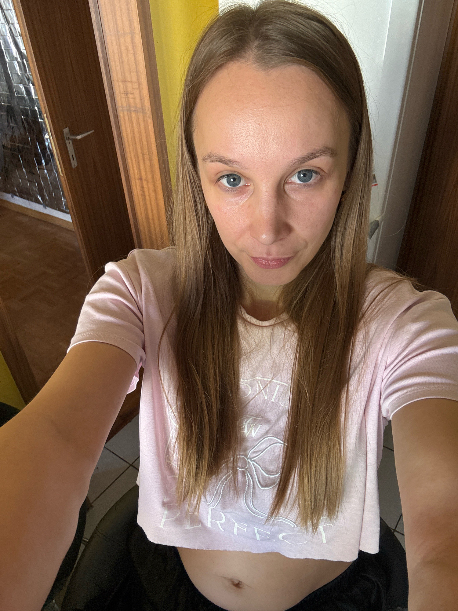 A young woman with long brown hair and blue eyes is pictured in a selfie. She is wearing a pink crop top with white lettering and a dark garment below. The background consists of wooden doors and a yellow wall, suggesting an indoor setting. The lighting is natural, and the overall atmosphere is casual. The image does not contain any information or visual cues that would allow for identification of the location as Überherrn, Germany.