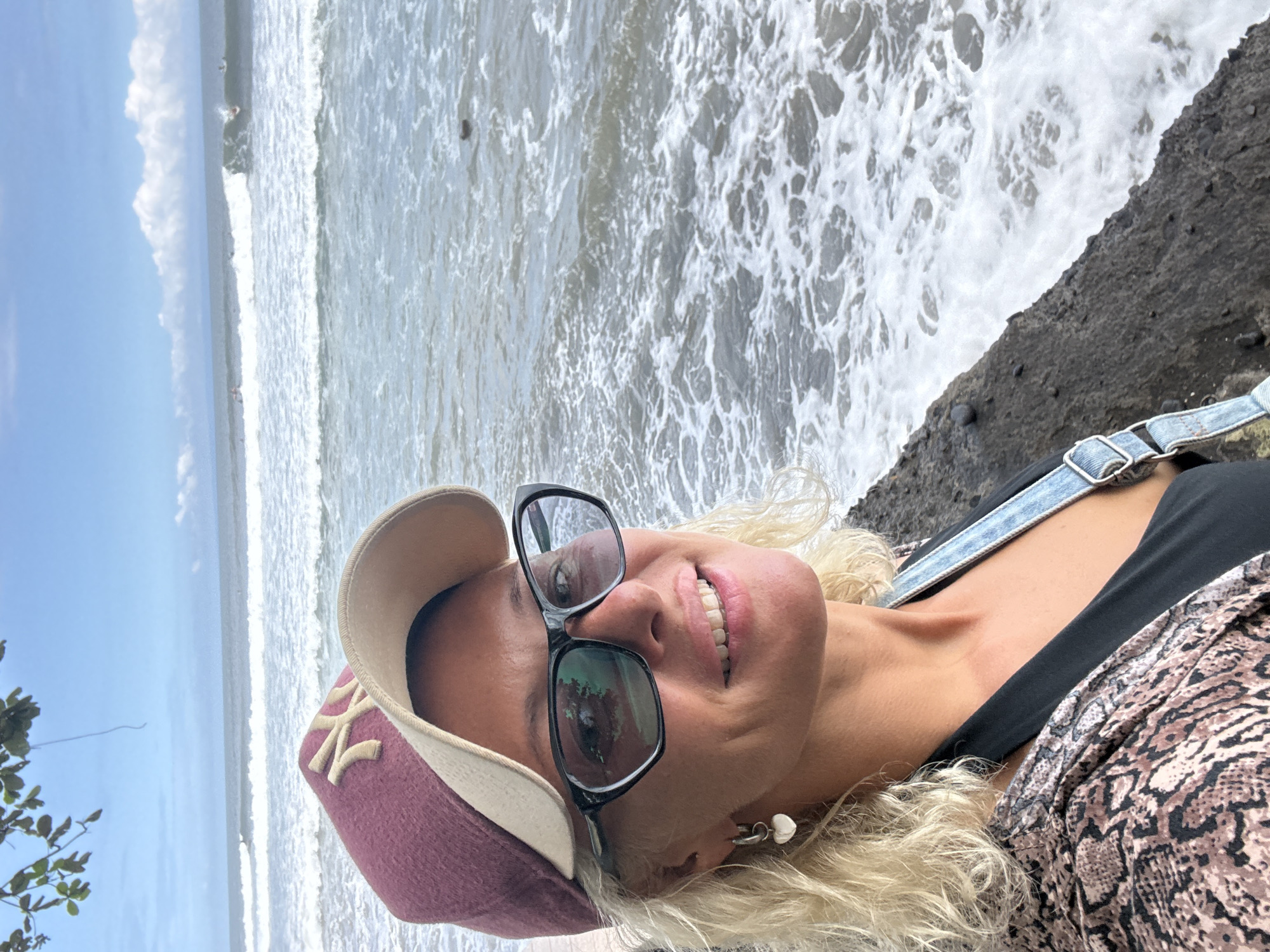A woman with blonde hair, wearing a burgundy baseball cap, black framed glasses, a black top, and a snakeskin patterned jacket, stands facing the camera. The backdrop is an ocean landscape, with crashing waves and a rocky ledge in the foreground. The location is Unknown, Unknown.