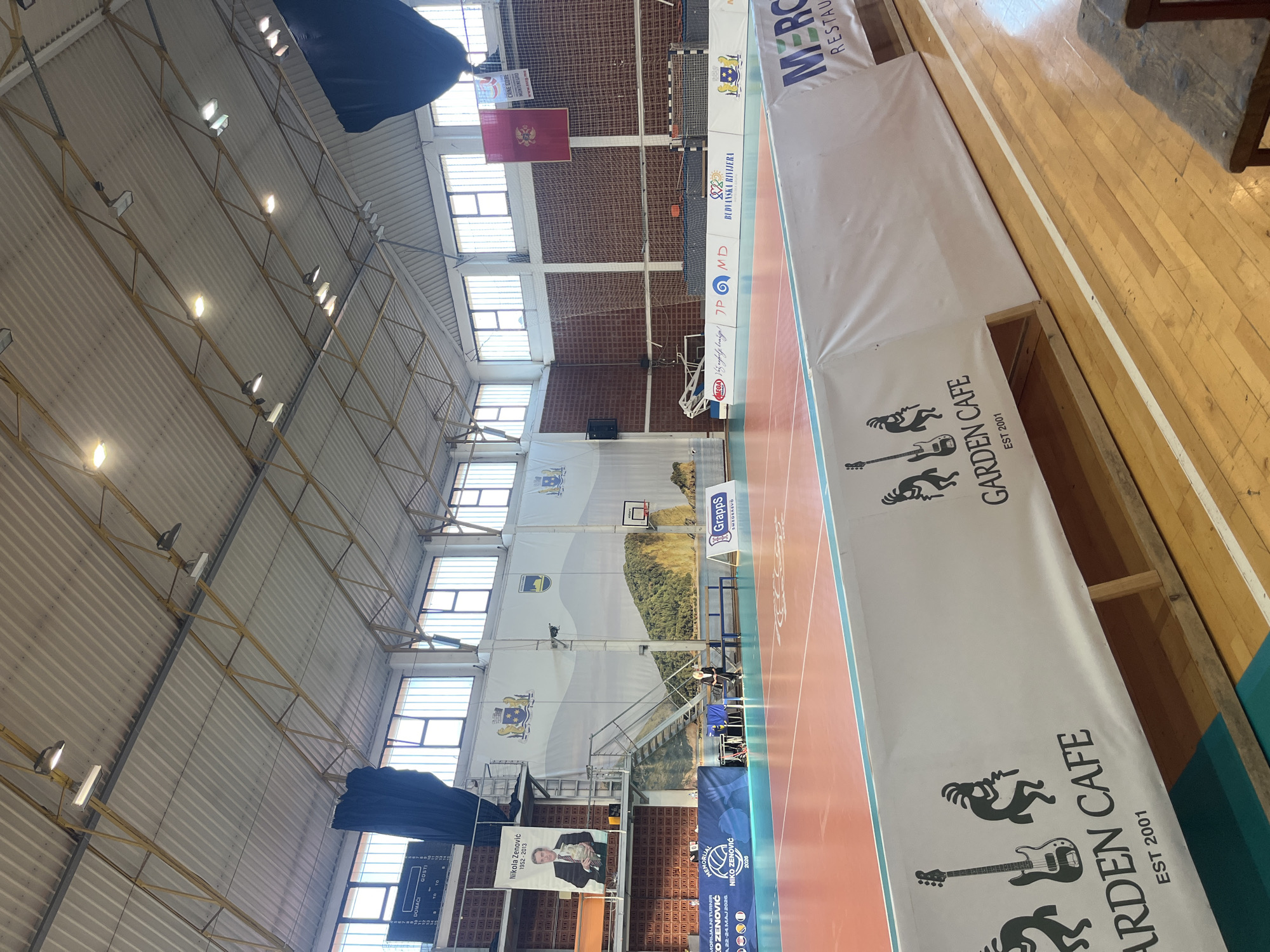 This is an indoor shot of a sports hall in Budva, Montenegro, likely used for basketball or handball. The hall has a brick wall at the far end, punctuated by several rows of windows. Banners are hung on the wall, including one displaying the flag of Montenegro. Another banner displays a graphic of a landscape, the Budva municipality seal, and a basketball hoop. Adjacent to it is another banner with the portrait of Nikola Zenović (1952-2013) and mentions "MORIJALNI TURNIR NIKO ZENOVIĆ" (Memorial Tournament Niko Zenović) along with dates (22-24.MAJ 2026). There is an electronic scoreboard showing scores. The court has red and white boundary lines. A barrier with "GARDEN CAFE EST 2001" branding is set up along the side of the court. The hall's ceiling is supported by yellow metal beams and has multiple light fixtures.