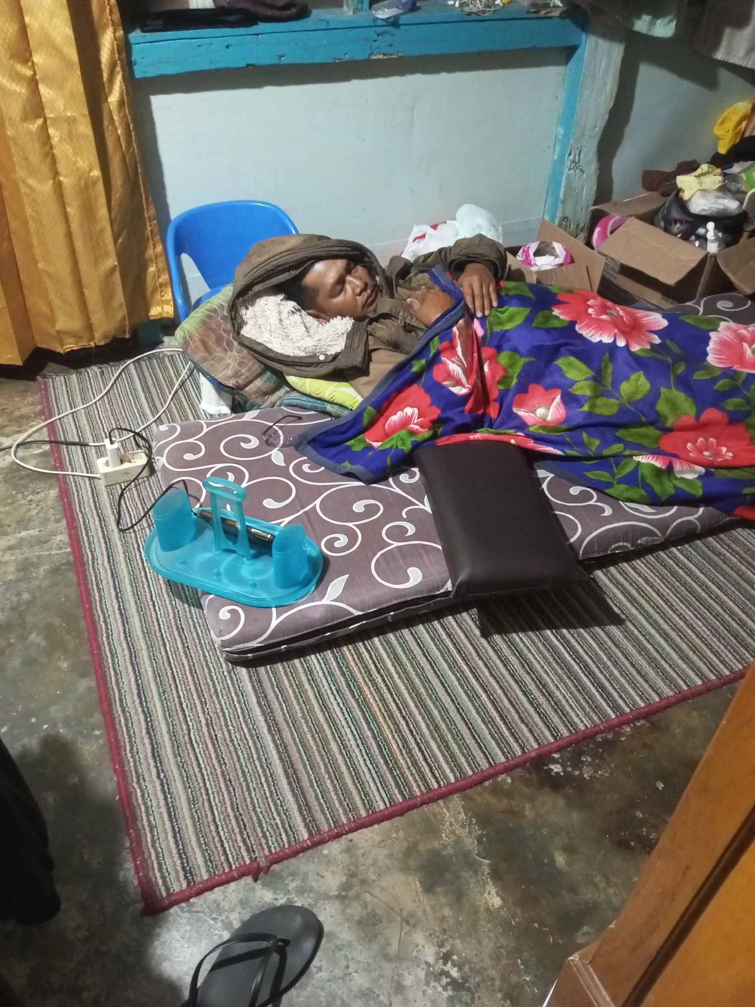 The image depicts a person sleeping on a mattress on the floor, covered by a floral blanket. The scene is likely indoors, possibly a room in Andongsili, Indonesia, characterized by simple furnishings and a somewhat cluttered environment. A yellow curtain hangs on the left, and a blue plastic chair is visible behind the sleeping person. Various objects are scattered around, including cardboard boxes, a power strip with a cord plugged in, and a turquoise plastic organizer with what appears to be a pen inside. A striped rug is laid out on the concrete floor, and a pair of black flip-flops are near the bottom right corner. The lighting appears to be artificial, casting shadows on the floor.