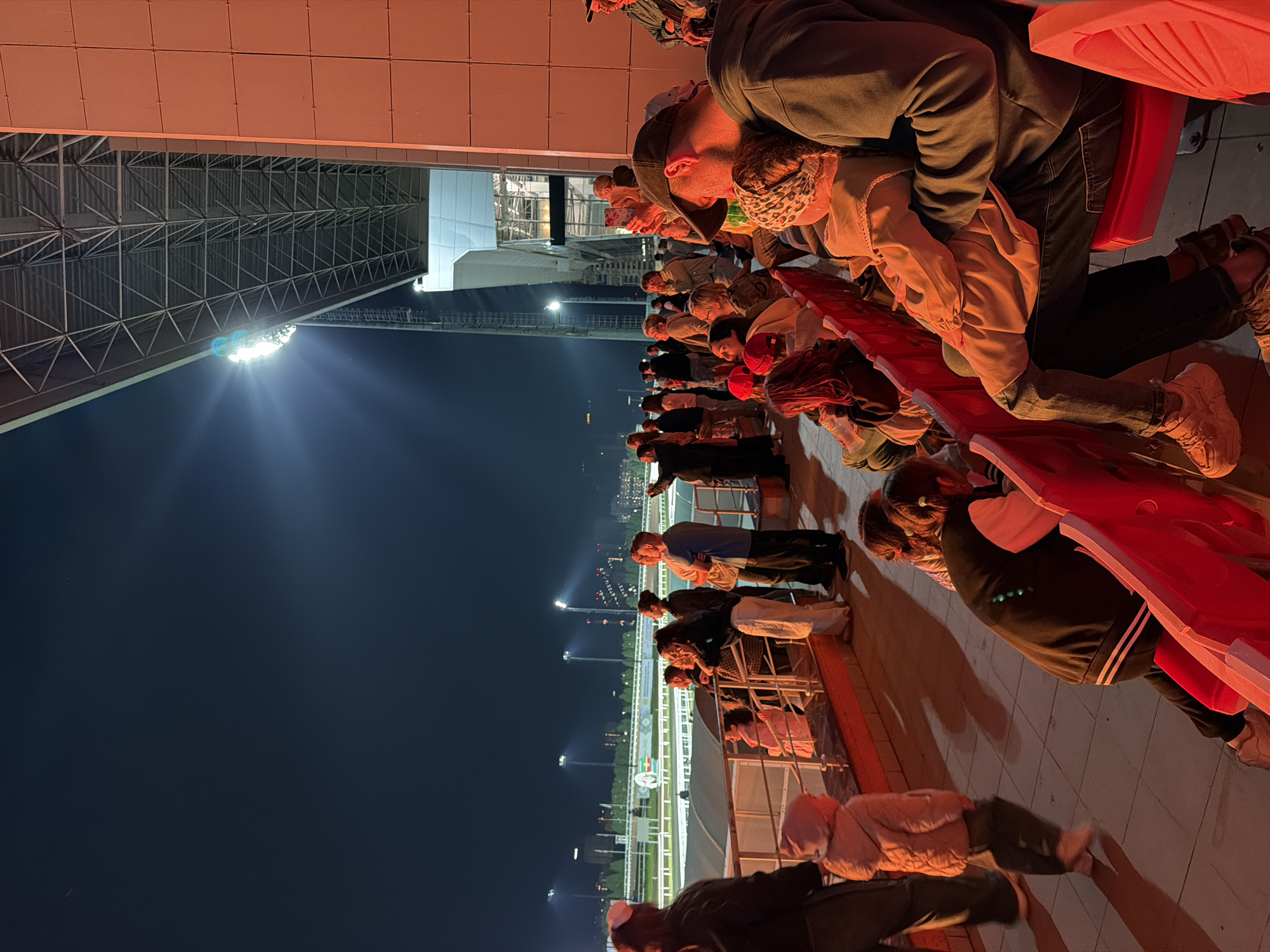 The image captures a nighttime scene at a horse racing track, with a crowd of spectators seated in illuminated bleachers. A bright light illuminates the track from above, while the surrounding area is cast in warm, reddish light, likely from stadium lighting. Spectators of various ages are visible, some standing and looking towards the track, while others are seated. A few children are also present, with one child in the foreground walking away from the camera. The architecture of the stadium is evident, with a metal roof structure and a concrete building. The overall atmosphere appears to be one of engagement and observation of the event.