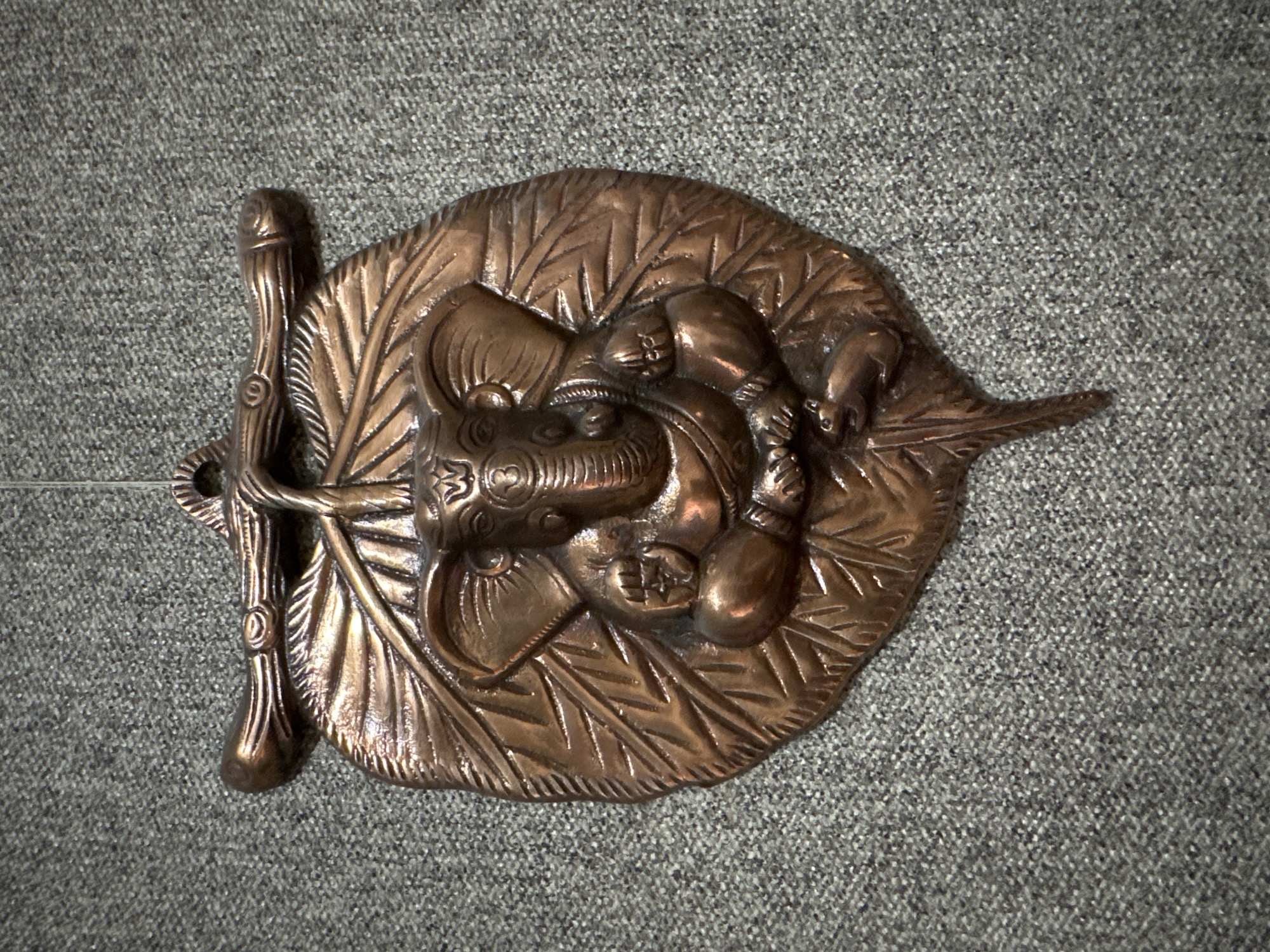 A copper wall hanging depicts the Hindu deity Ganesha seated on a leaf, with a small mouse at his feet. The hanging is suspended from a thin wire against a textured gray background.
