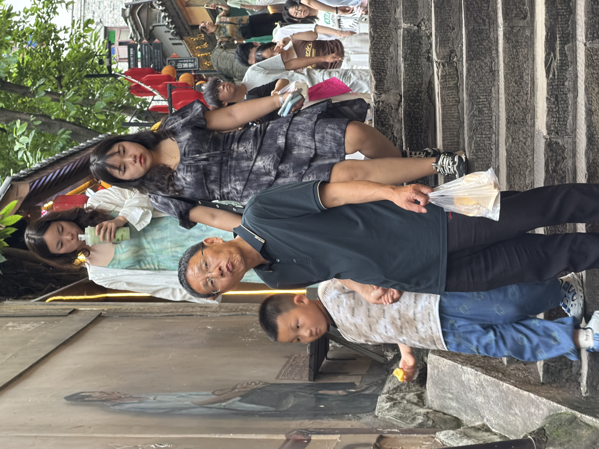 This image depicts a group of people on a stone staircase in Yuzhong District, China. The central figures are a man and a young boy. The man, wearing a dark polo shirt and glasses, is making a puckered face towards the camera while holding a plastic bag with food. The boy, in a patterned t-shirt and blue pants, is holding a piece of food in his left hand and standing on a lower step. Behind them, several other individuals are visible on the stairs, including a woman in a white shirt and a teal dress holding a drink, and another woman in a dark, ruffled dress. The staircase is flanked by textured walls, and the background shows hints of buildings and greenery, characteristic of Yuzhong District's hilly terrain and traditional architecture. Red lanterns are visible hanging from an overhang to the right, suggesting a festive or commercial area. The overall scene captures a moment of everyday life on a busy street in China.