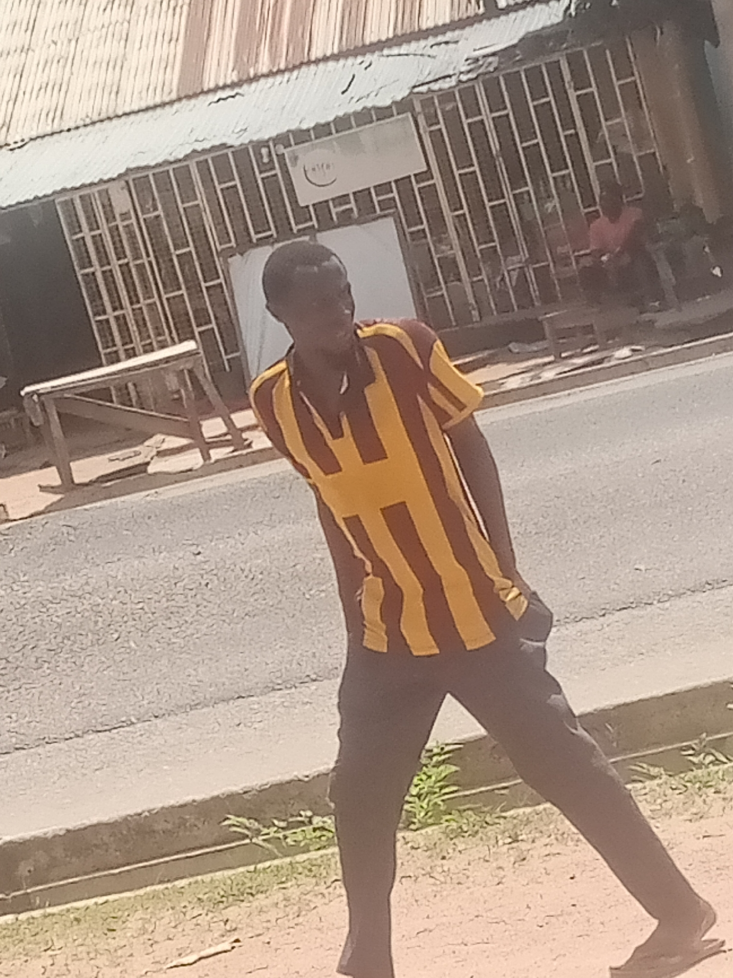 A young man wearing a striped brown and gold shirt and dark pants stands on a street in Jalingo, Nigeria. He is looking to his right, with his hands in his pockets, and appears to be walking. The street is paved and there is a gutter with some greenery on the side. Behind him, there is a building with a corrugated metal roof and barred windows. A table or stand is visible in front of the building. In the background, another person can be seen sitting on a stool in front of the building. The scene appears to be in daylight.