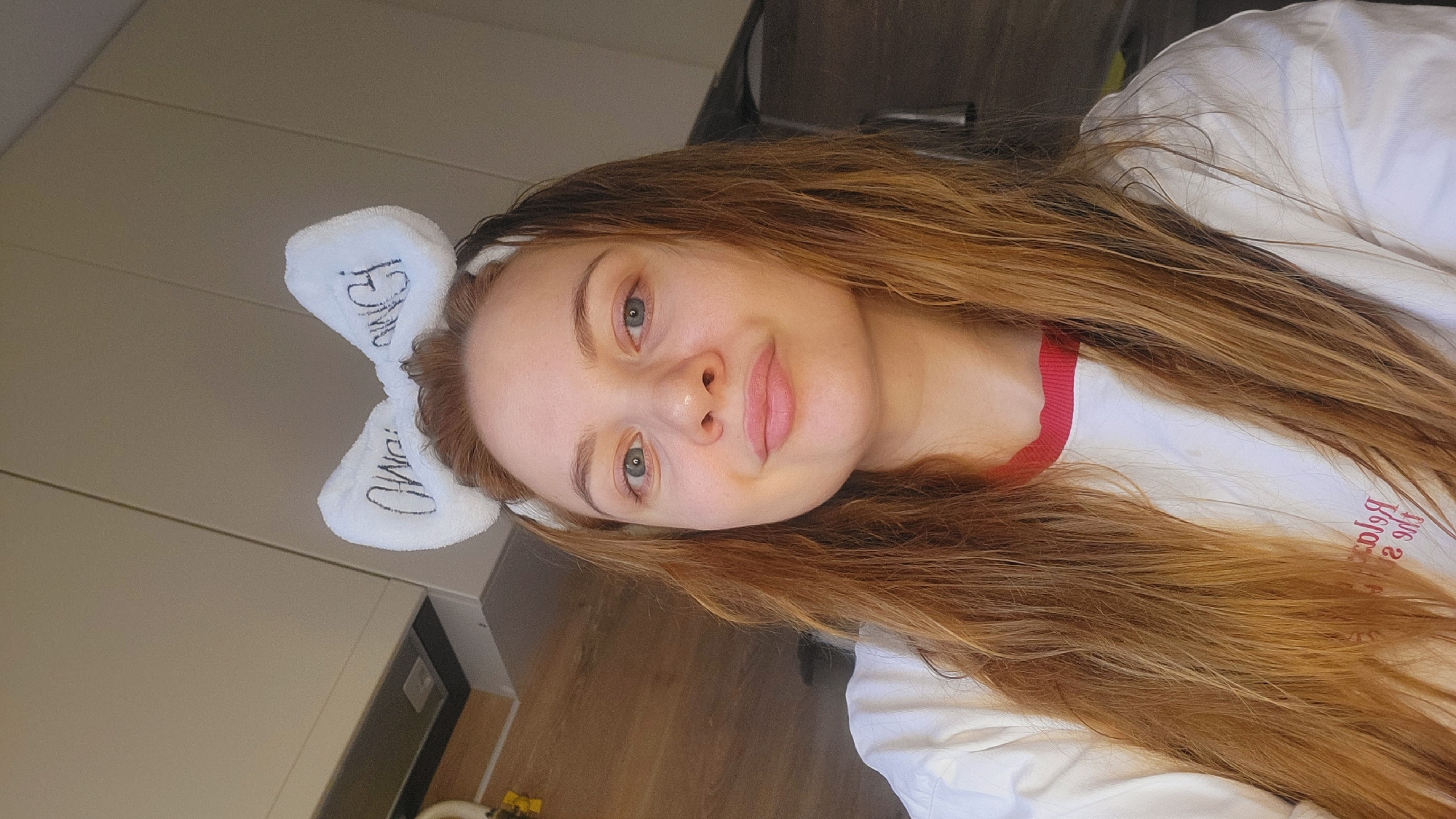 A woman with long brown hair wearing a white top with a red collar and a white headband with bunny ears is posing for a selfie. The words "For MD" are written in black on each ear of the headband. The background is a combination of neutral and bright lighting on the upper and lower portions, respectively. It seems the photo was taken in a domestic environment, potentially a bathroom or bedroom. There is no definitive indication of Chaikovsky, Russia in the image.