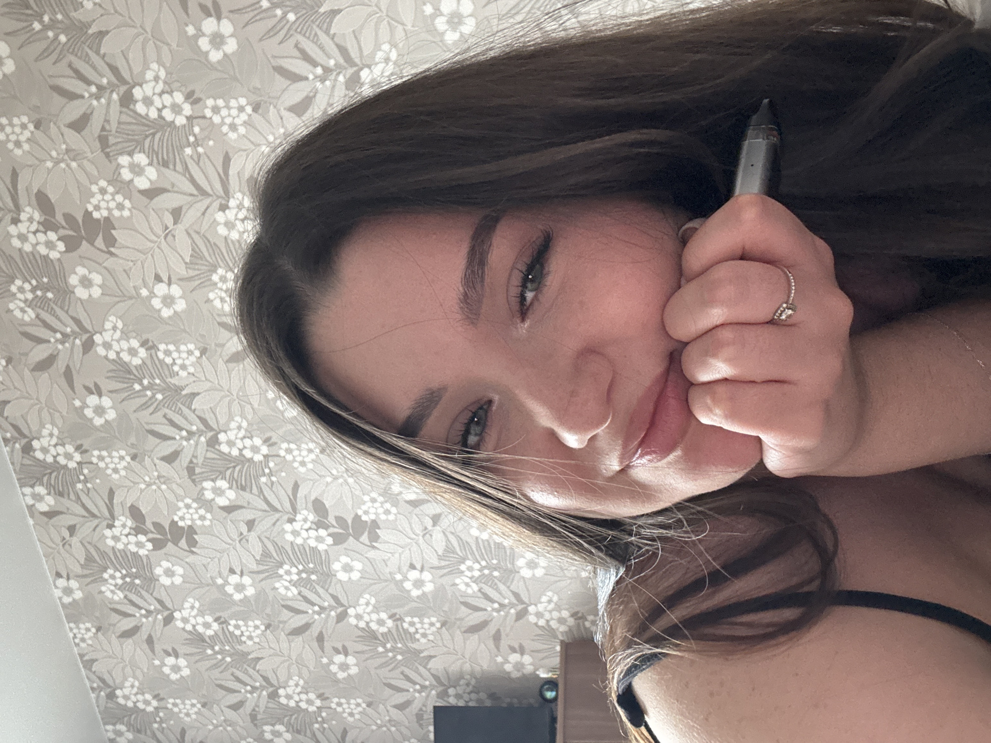 A woman with long, brown hair and fair skin is holding a vape pen near her face, with her hand resting against her cheek. She is wearing a ring on her finger. The background is a grey floral patterned wallpaper. A black tank top is visible on her shoulder. It's not possible to determine the location in Perm, Russia from the image.