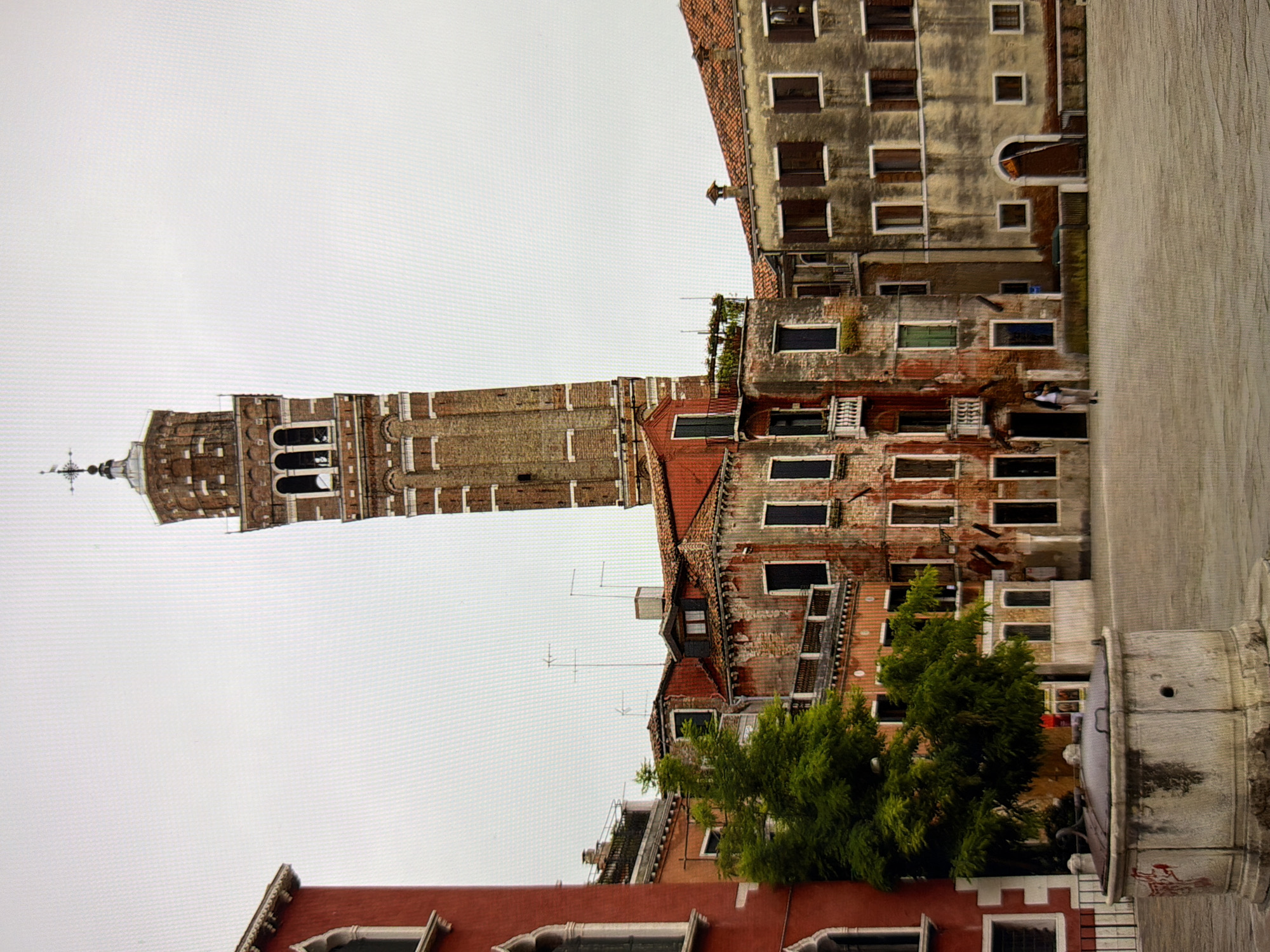 This image depicts a square in Venice, Italy, dominated by a tall, narrow brick bell tower with arched windows and a cross at its apex. The bell tower is surrounded by several aged, multi-story buildings with terracotta tiled roofs, weathered facades, and numerous windows. A large, old stone well with a metal lid is prominently featured in the foreground on the left side of the square. A lush green tree stands behind the well. Two individuals are walking in the middle ground of the square, and the sky is overcast and pale. The architecture and atmosphere are characteristic of Venice, Italy, not Kyiv, Ukraine.