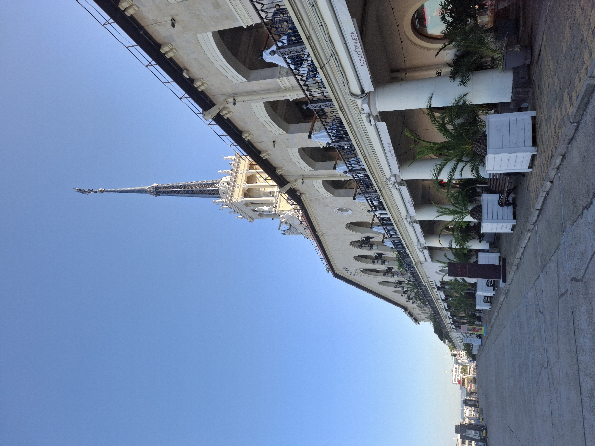 The image captures a wide shot of the Sochi Sea Port in Sochi, Russia. The architecture is distinctive, with a light-colored facade featuring a series of arched walkways and a tall, spired tower. The building is adorned with balconies, classical columns, and decorative elements. In front of the building, white planters with palm trees are arranged along the sidewalk. The ground in the foreground is a concrete walkway. The sky above is a clear, bright blue. Signs for "BOSCO RIVIERA" are visible above the ground-floor businesses. There are no people clearly visible in the frame. The scene appears to be set during daylight, with a clear sky and good visibility.
