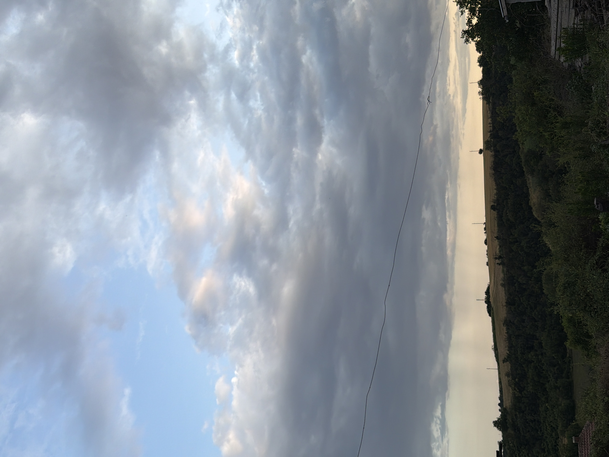 The image shows a view of rolling hills covered with trees and fields under a dramatic cloudy sky. In the foreground, there is dense green vegetation and what appears to be part of a building on the right. A dark wire runs horizontally across the middle of the frame, bisecting the sky and the landscape. In the distance, atop the hills, there are a few scattered structures, possibly including utility poles and a solitary tree. There is a visible rain shower in the far distance, indicated by streaks of precipitation against the lighter sky. The overall scene suggests an overcast or approaching storm over a rural or semi-rural area. This image was taken in the city of Nikopol, Bulgaria.